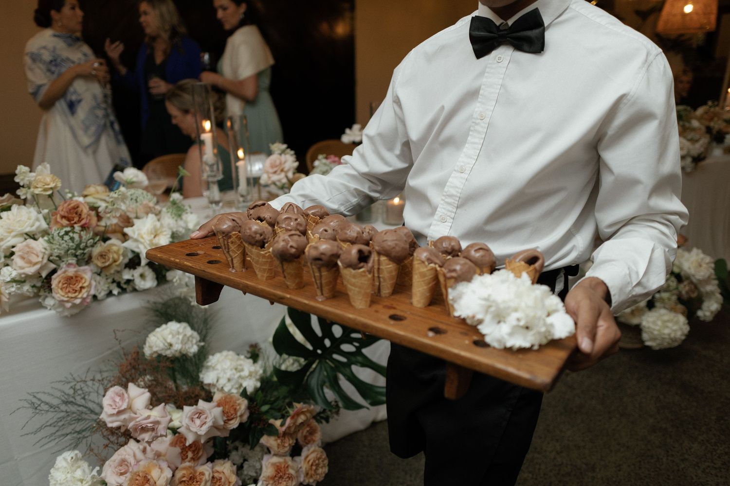 Waiter in bow tie holding wooden tray with ice cream cones at elegant wedding reception with floral arrangements.