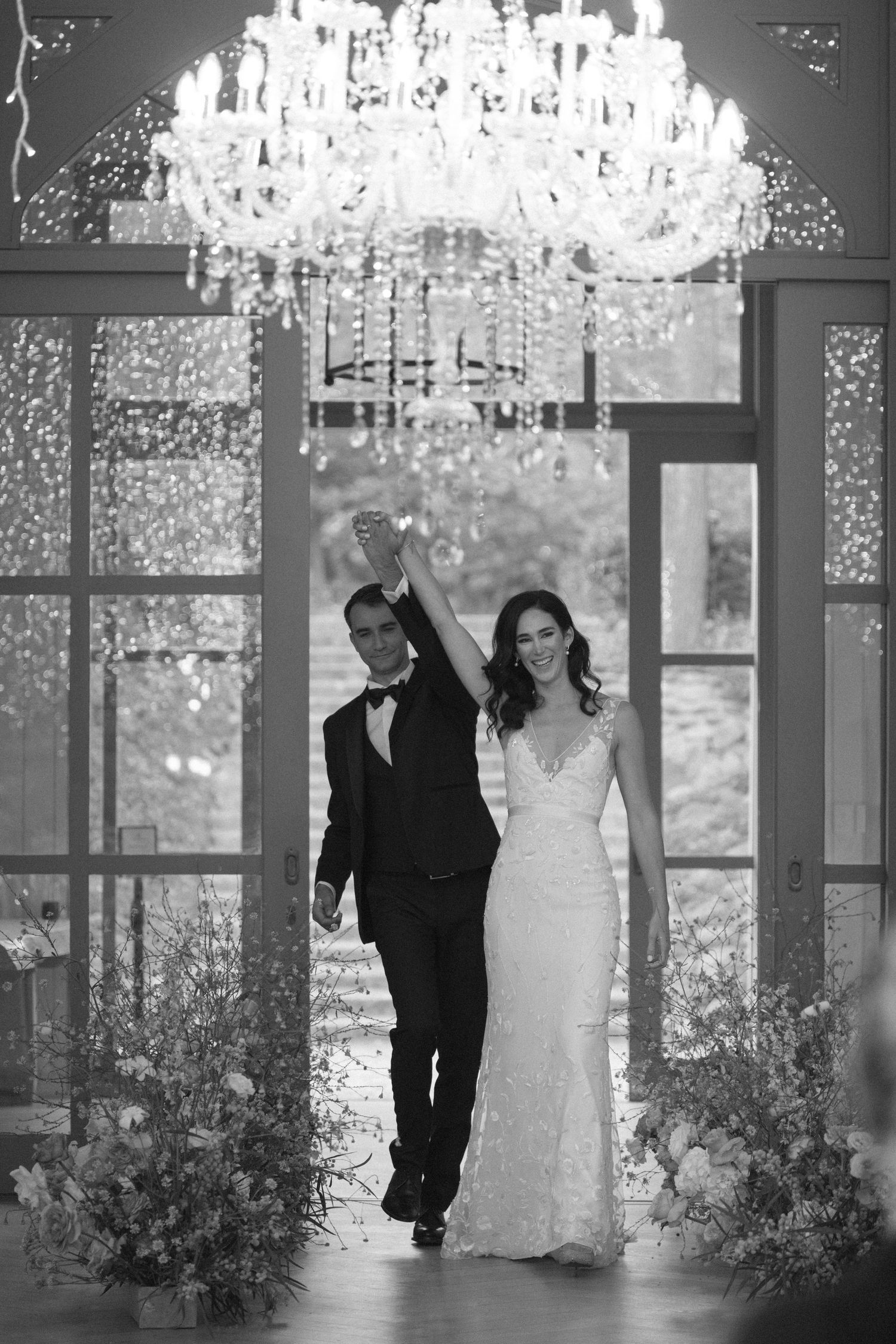 Black and white photo of newlyweds making grand entrance under crystal chandelier.