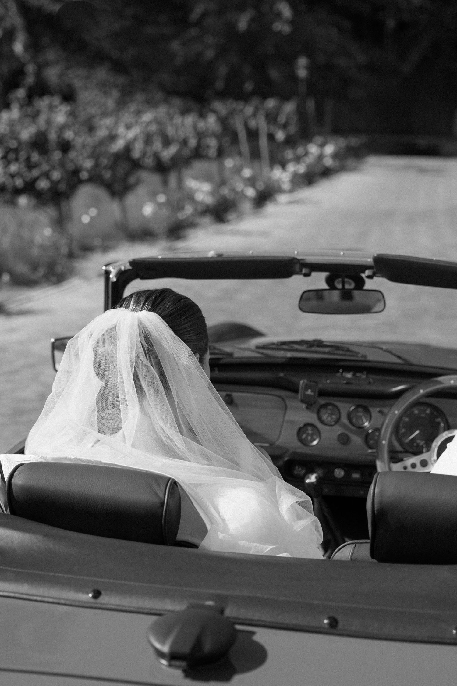 Black and white photo of a bride in veil sitting in a vintage convertible car on her wedding day.