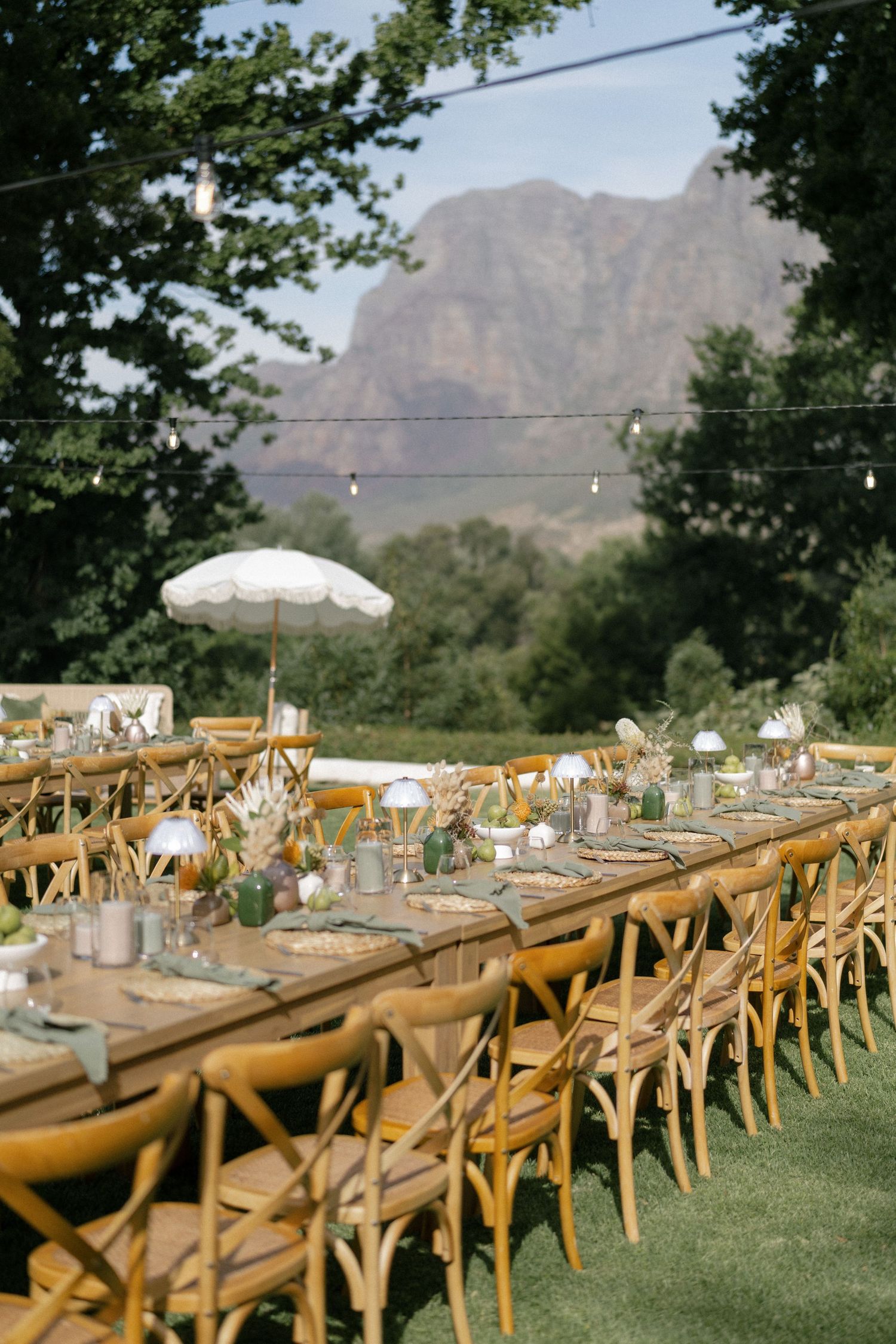 Elegant outdoor wedding reception with long wooden tables, cross-back chairs, and mountain backdrop under string lights.