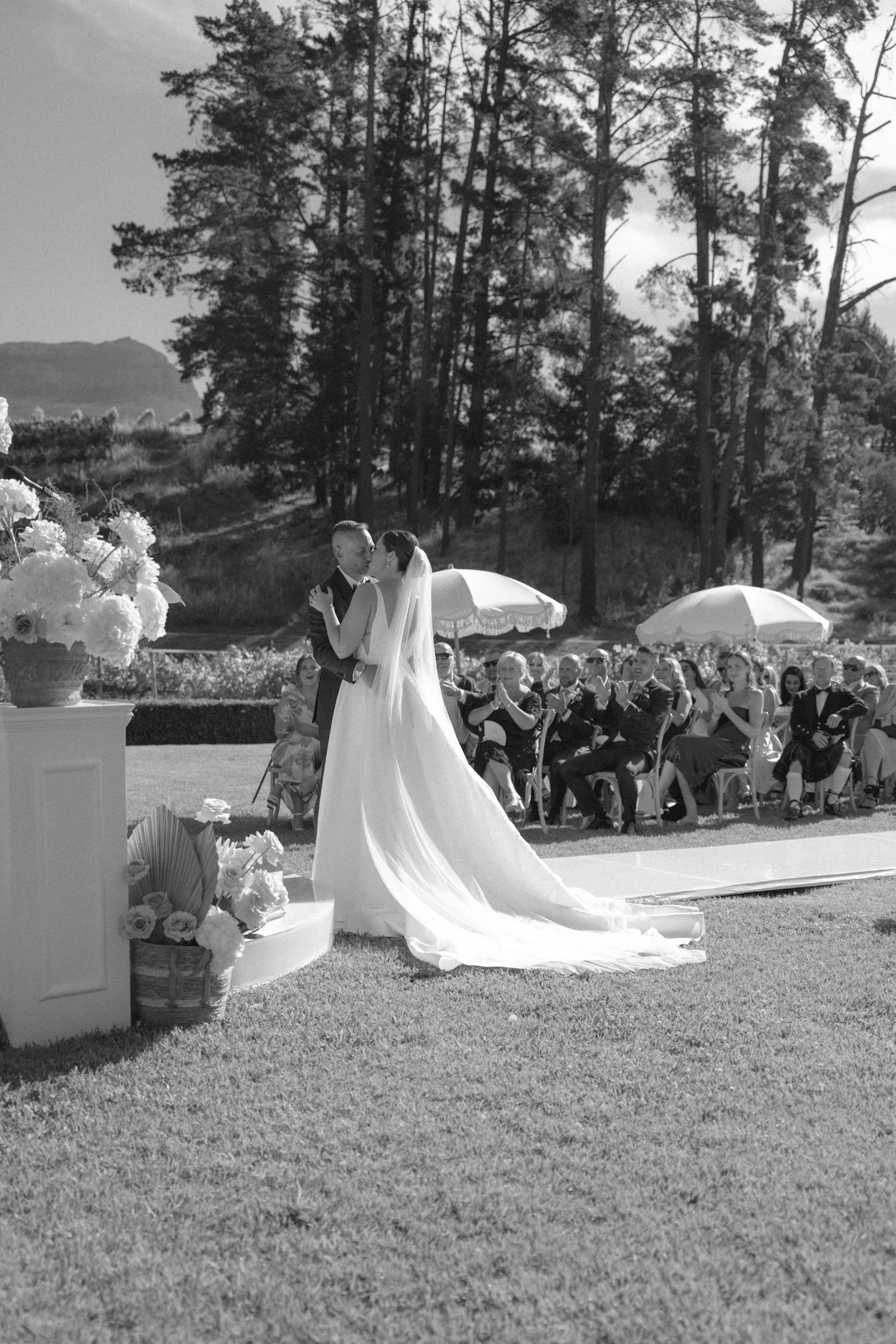 Black and white photo of bride and groom sharing first kiss at outdoor wedding ceremony with guests seated behind them.