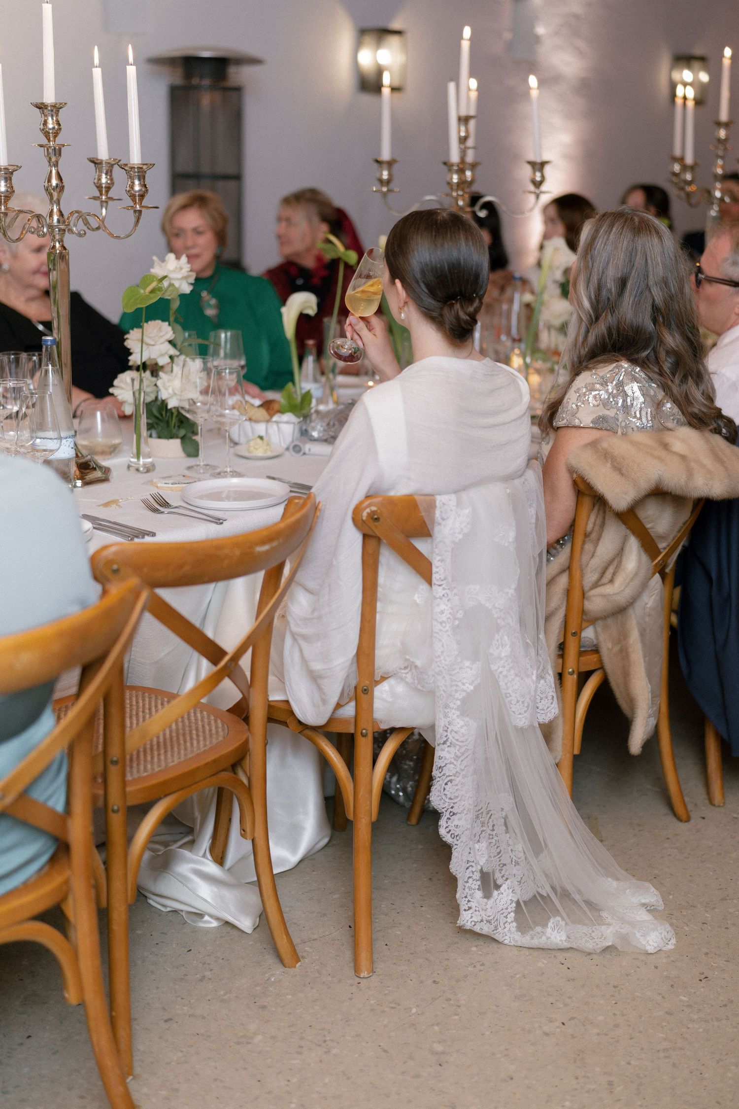 Bride in lace wedding gown seated at elegant candlelit reception dinner table with guests in formal attire.