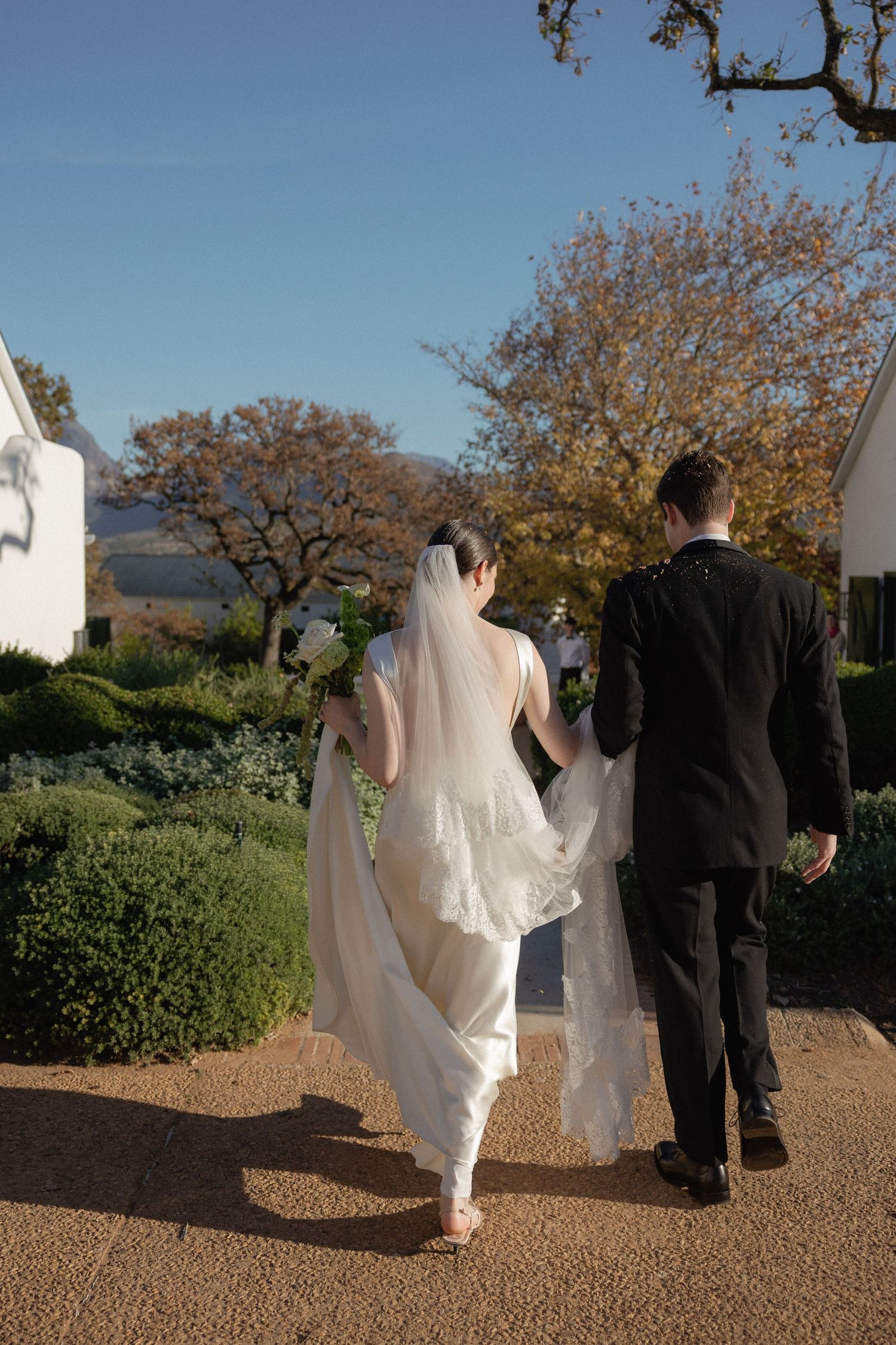 Bride in white gown with flowing veil and groom in black suit walk hand in hand through manicured garden path.