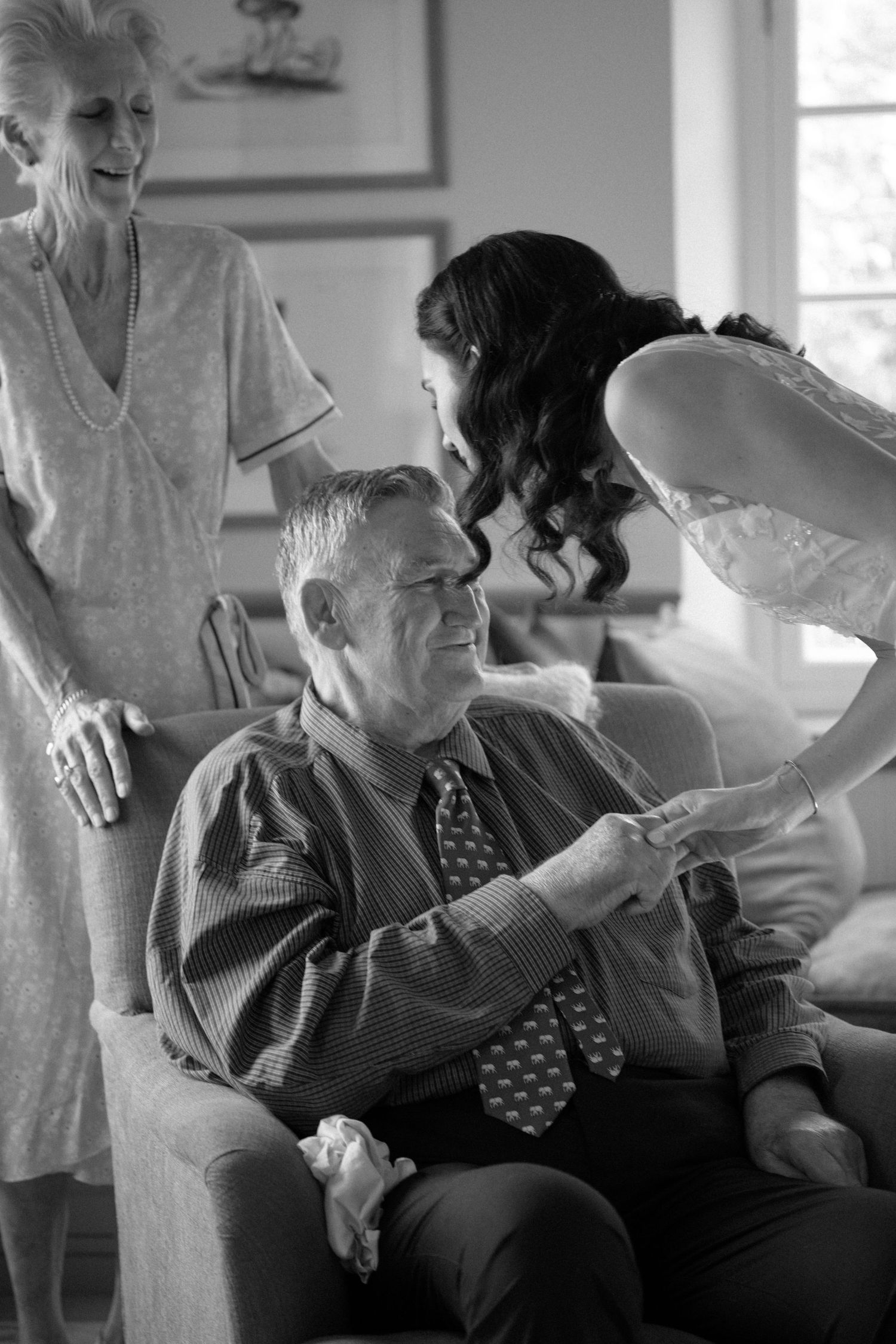 Black and white photo of a bride in a beaded gown sharing a tender moment with seated elderly grandparents indoors.