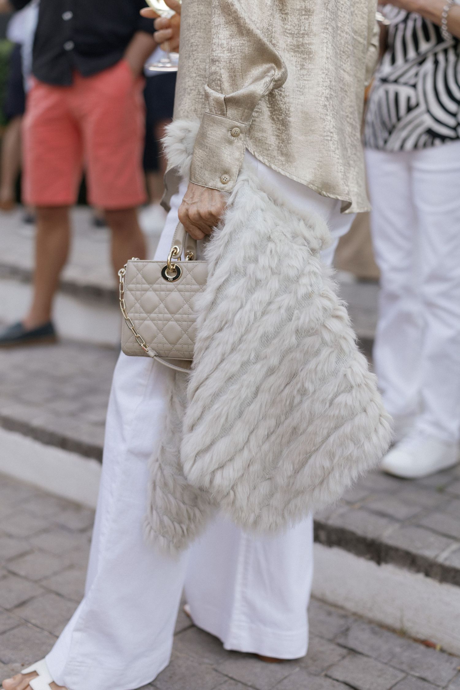 Woman in white wide-leg pants holding a quilted mini bag, wearing a cream fur vest and metallic jacket outdoors.