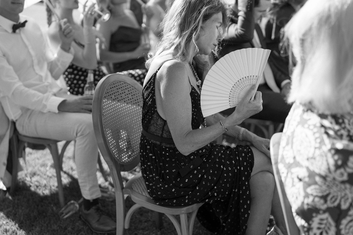 Woman sitting in chair at outdoor event, holding white hand fan, wearing polka dot dress, black and white photo.