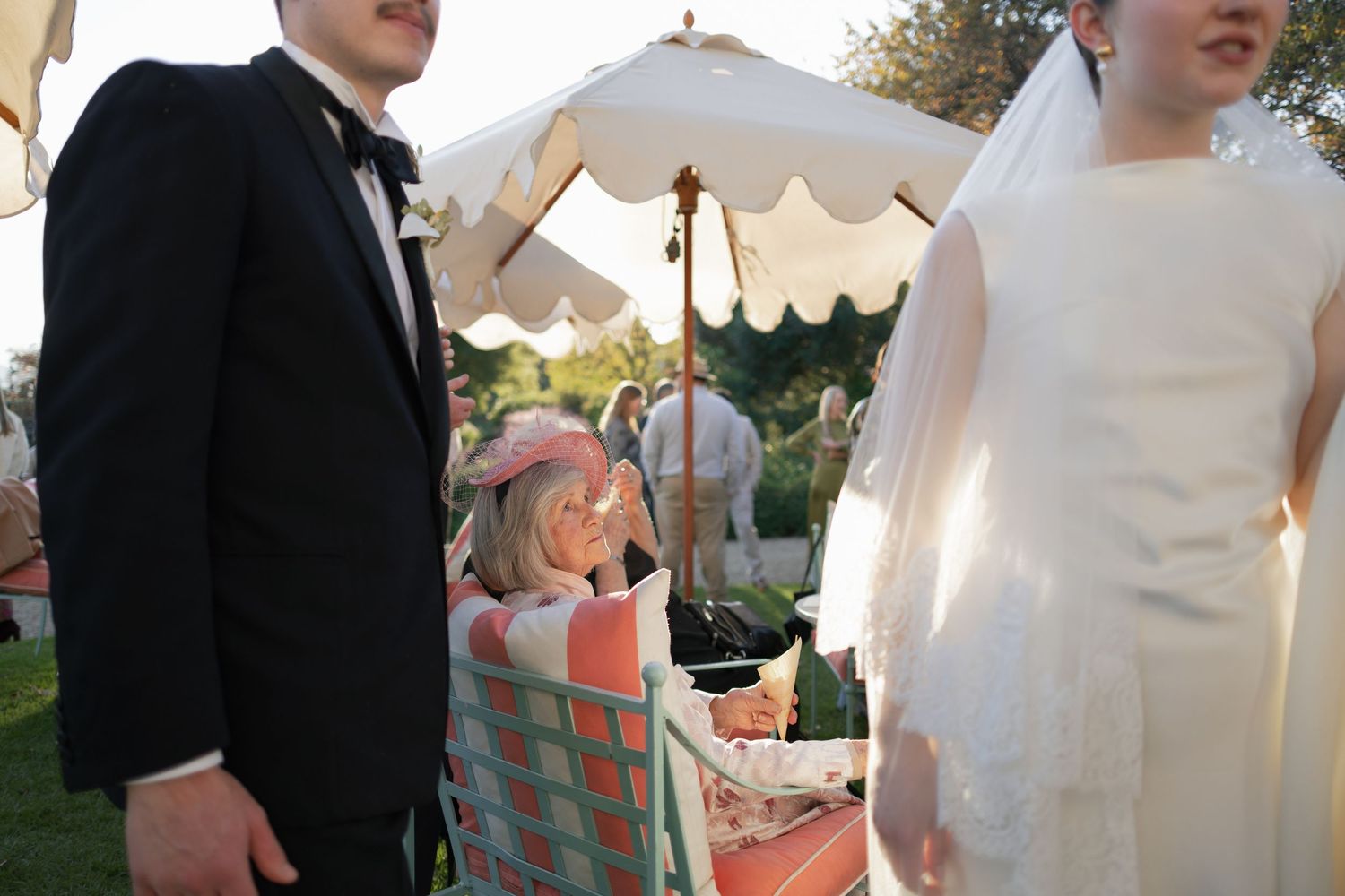 Bride in white veil and groom in tuxedo standing near a child at an outdoor wedding reception.