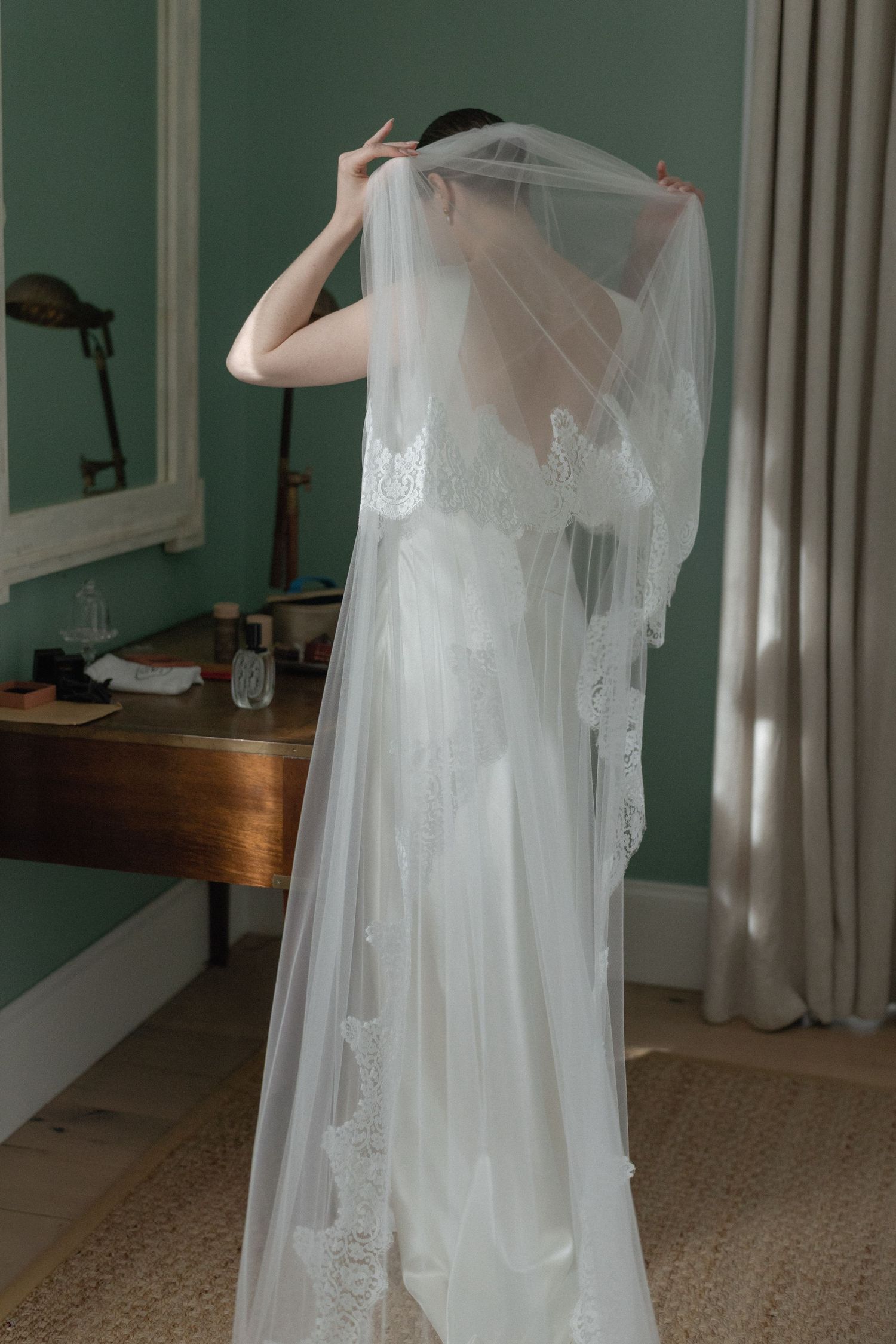 Bride in backless satin gown adjusts long lace-trimmed veil before mirror.