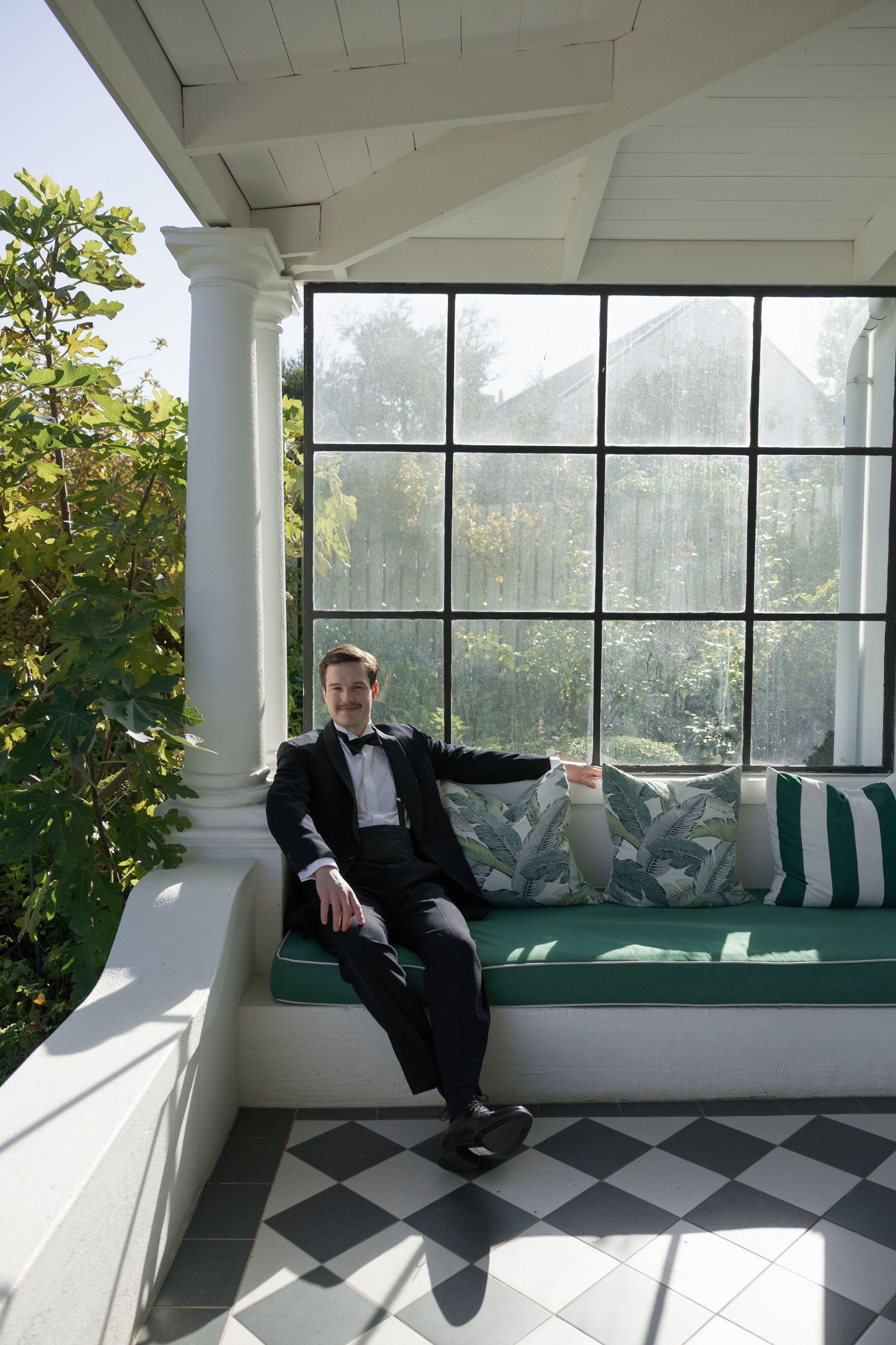 Dapper groom in black tuxedo relaxes on a cushioned porch bench with checkered floor and large windows.