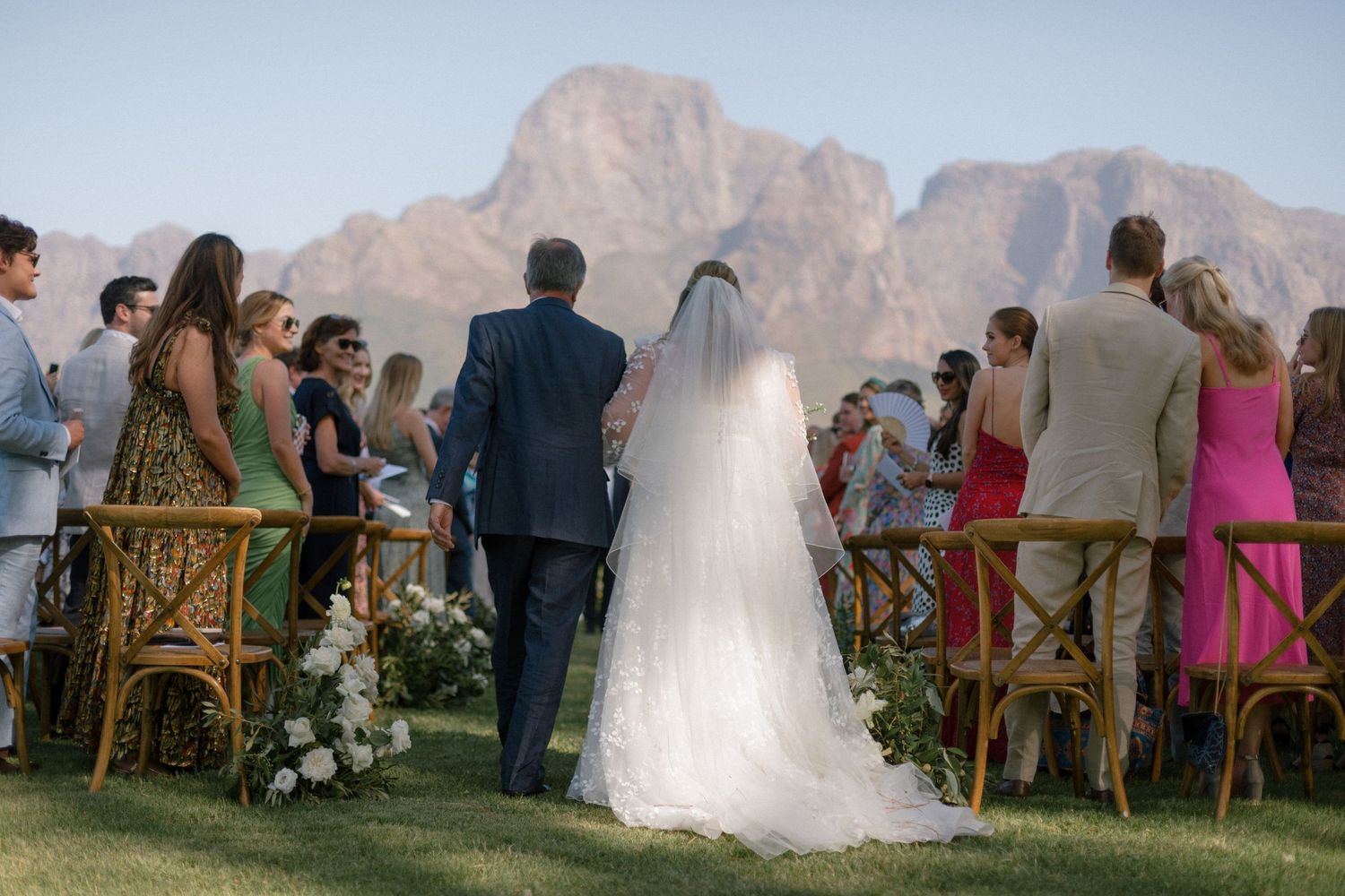 Bride walking down outdoor aisle with escort toward mountain backdrop at scenic garden wedding ceremony.