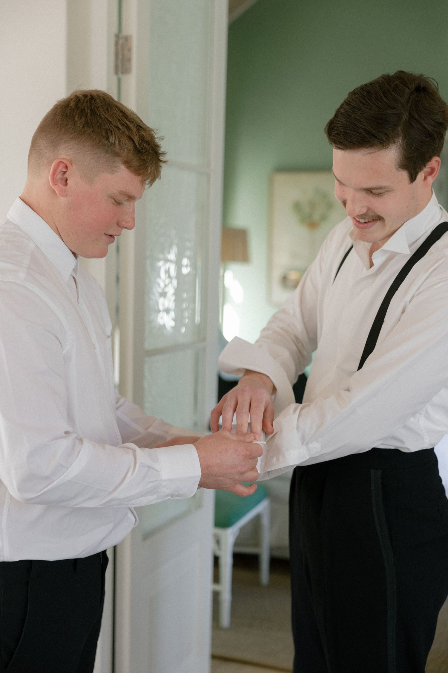 Groomsman helping smiling groom fasten cufflinks while wearing white shirts and black suspenders.