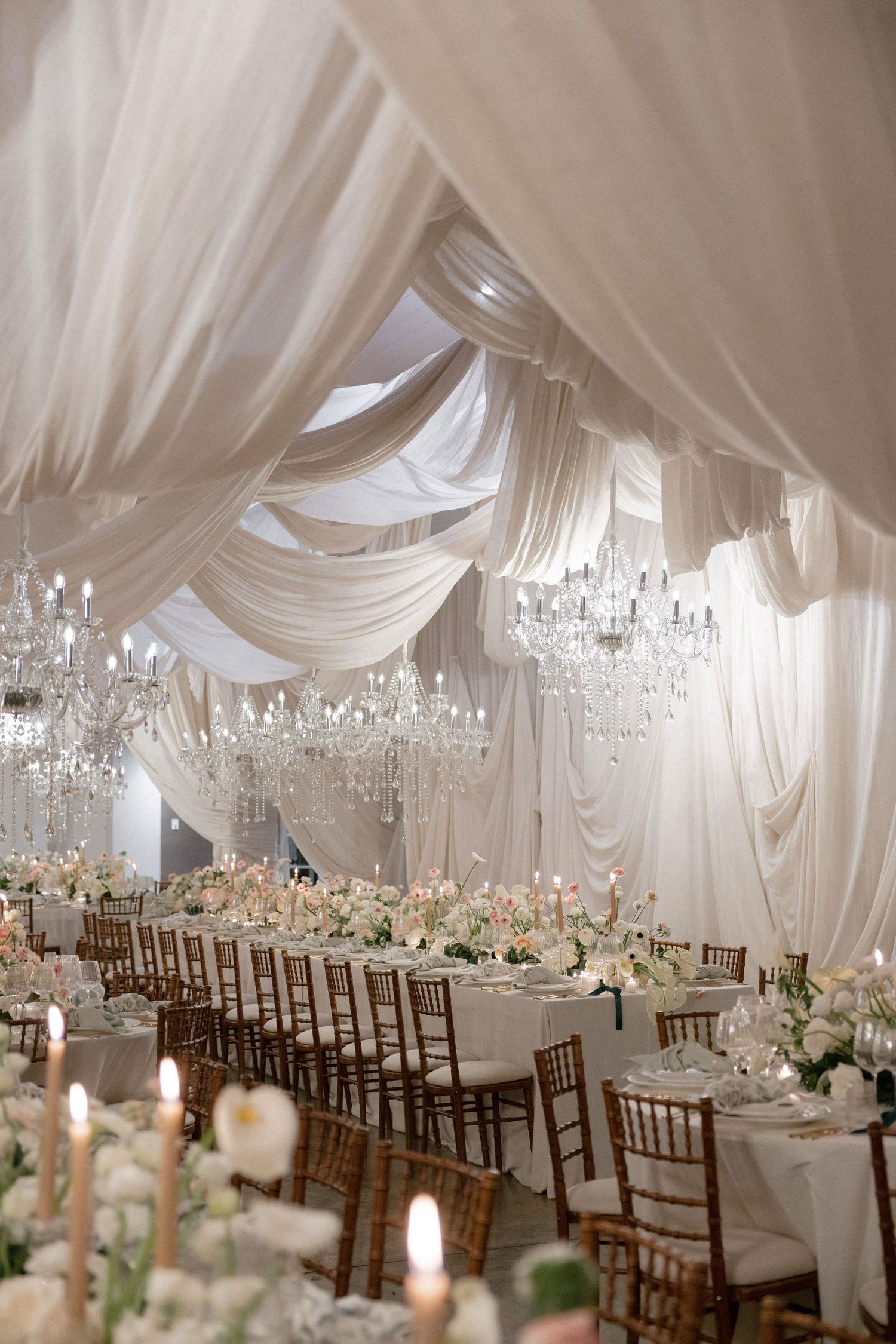 Luxurious wedding reception hall with white draped ceiling, crystal chandeliers, and floral centerpieces.