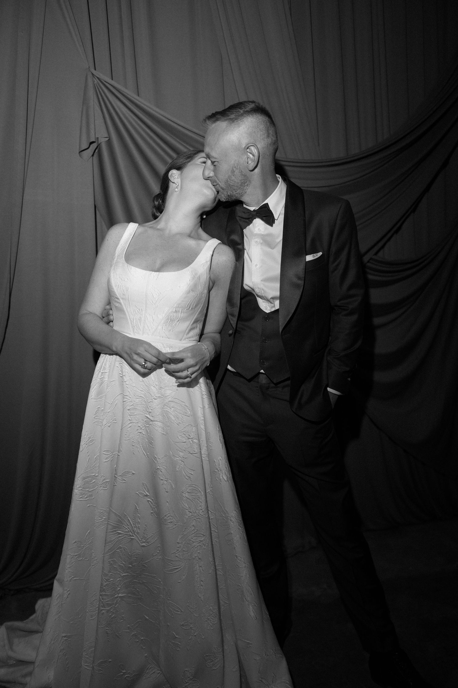 Bride and groom share a tender kiss in black and white portrait against draped fabric backdrop.