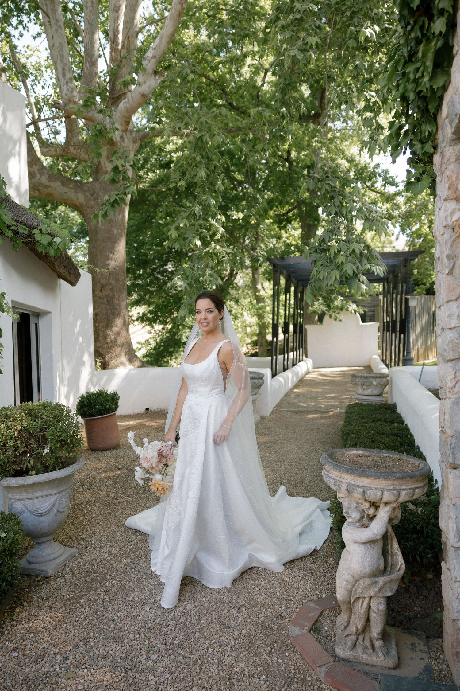 Bride in white gown with veil poses in a sunlit garden courtyard with stone statues and lush green trees.
