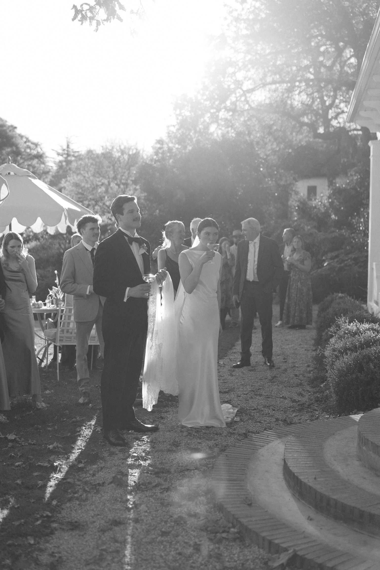 Black and white photo of bride and groom standing together at sunlit outdoor wedding reception with guests.