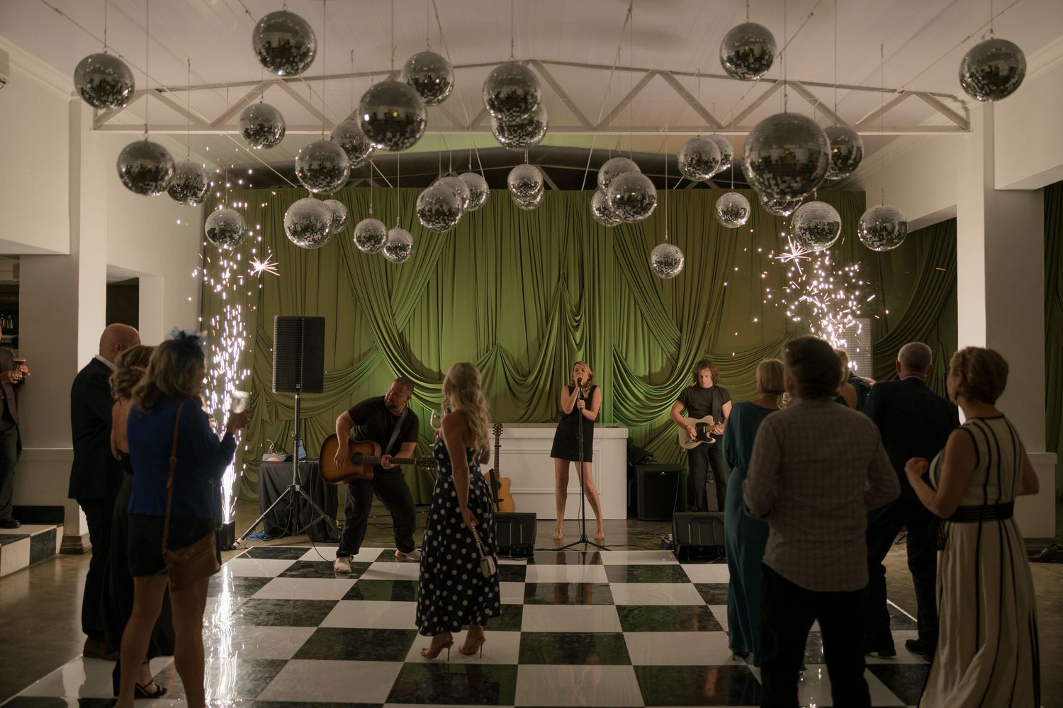 Elegant event venue with disco balls, green drapes, live band, and guests on a black-and-white checkered dance floor.