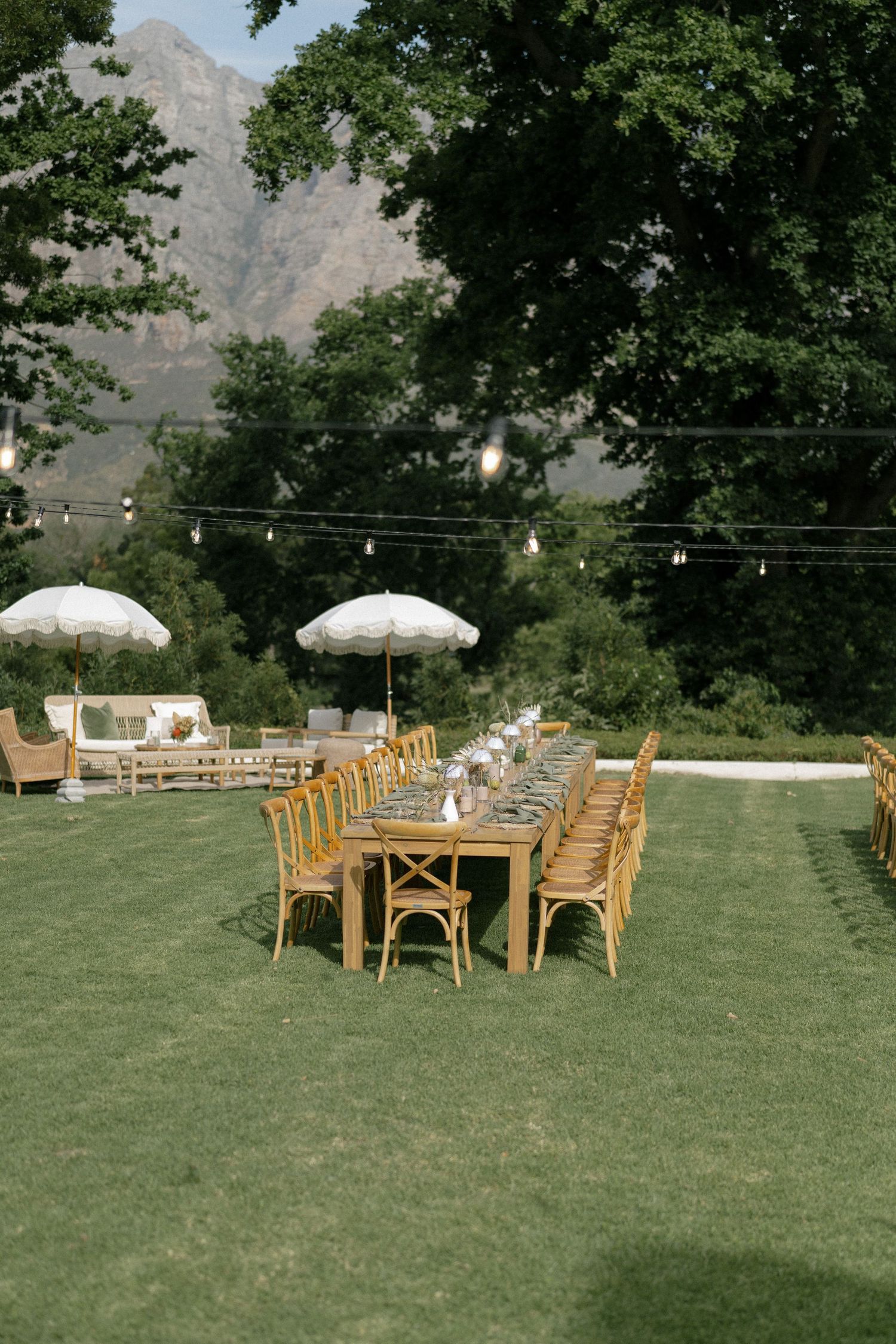 Elegant outdoor wedding reception setup with wooden chairs and long table on green lawn, framed by mountains and string lights.