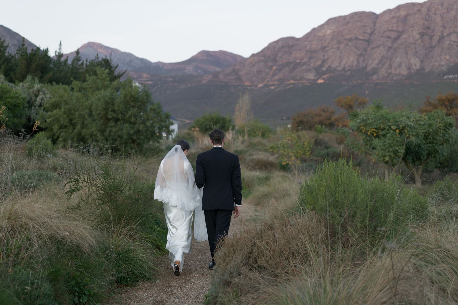 Bride and groom walking hand in hand along a scenic mountain trail at dusk.