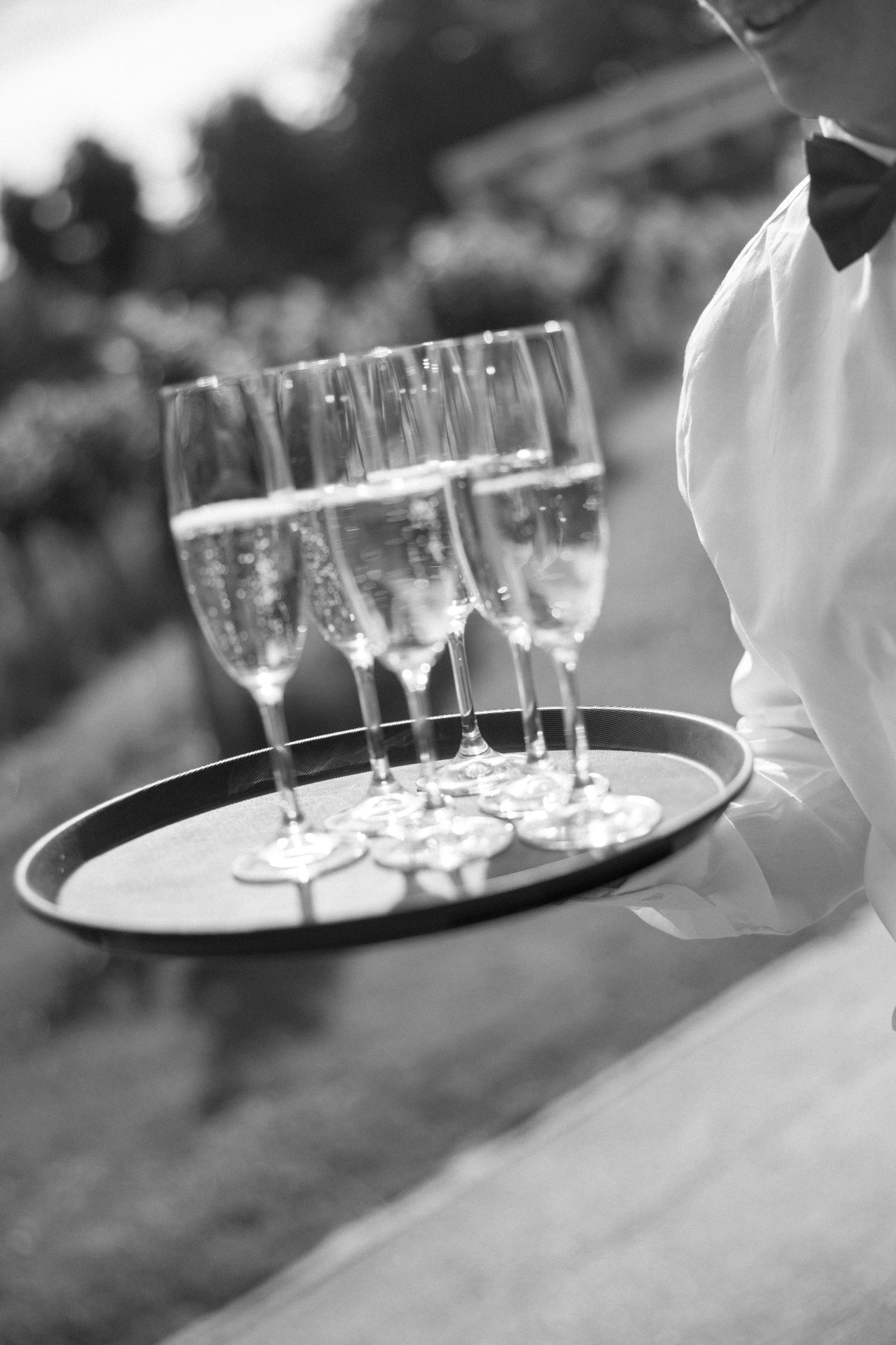Black and white photo of a waiter holding a tray of champagne flutes and appetizers outdoors.