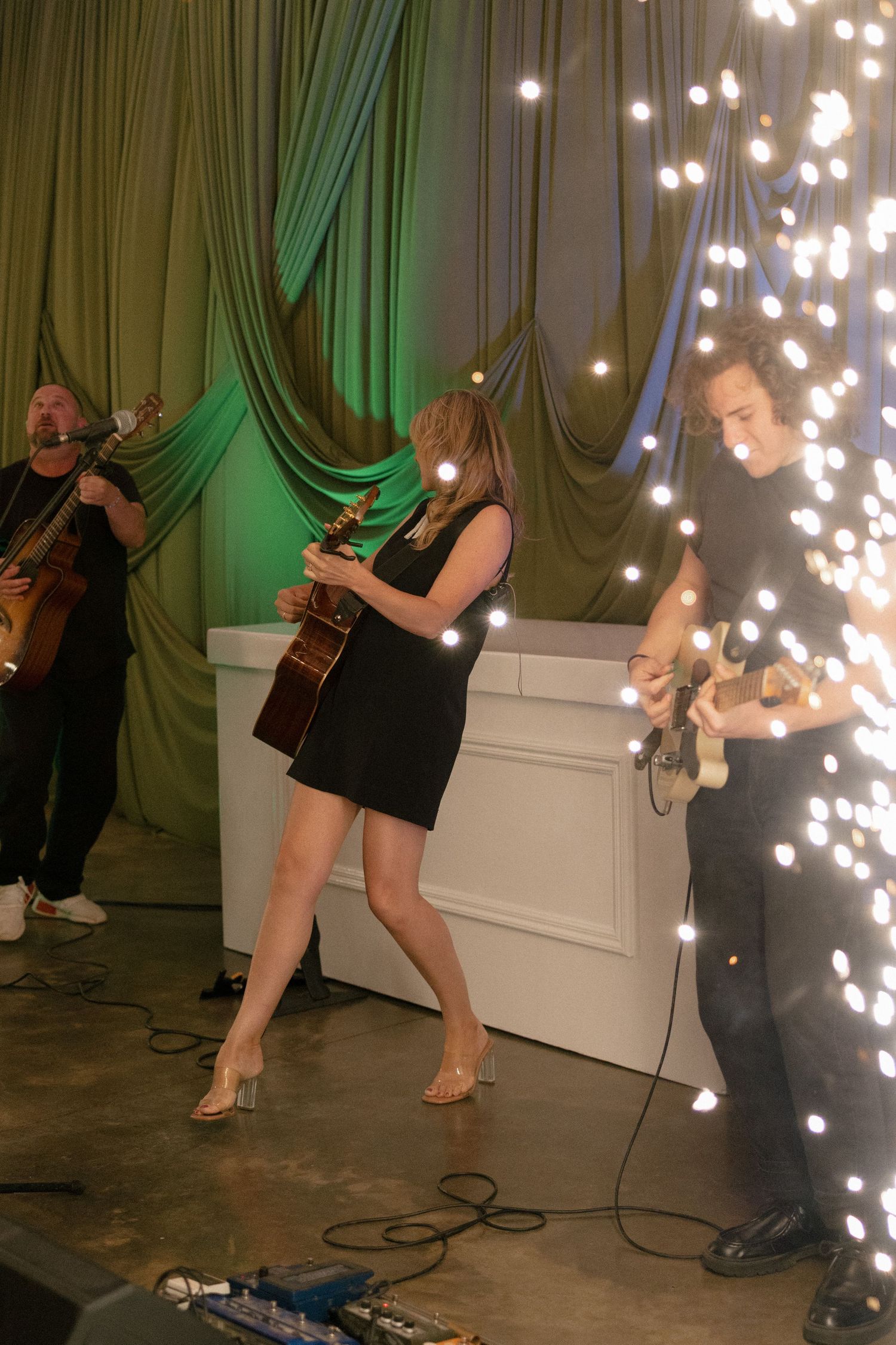Live band performance with three musicians playing guitars on stage with green drapes and string lights backdrop.