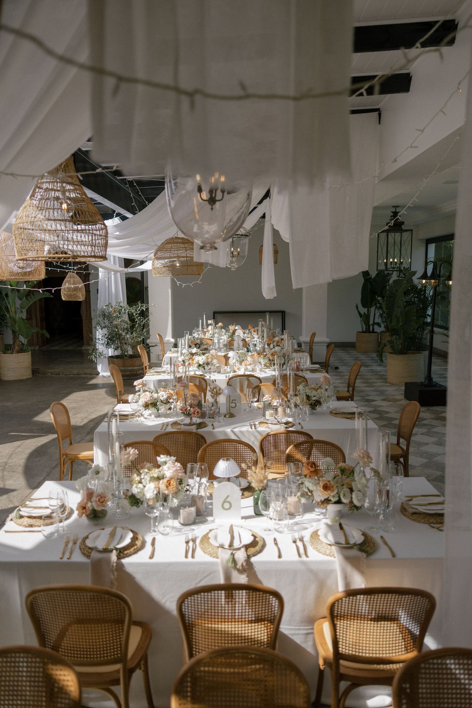 Elegant wedding reception table with white linens, floral centerpieces, gold accents, rattan chairs, and woven pendant lights.