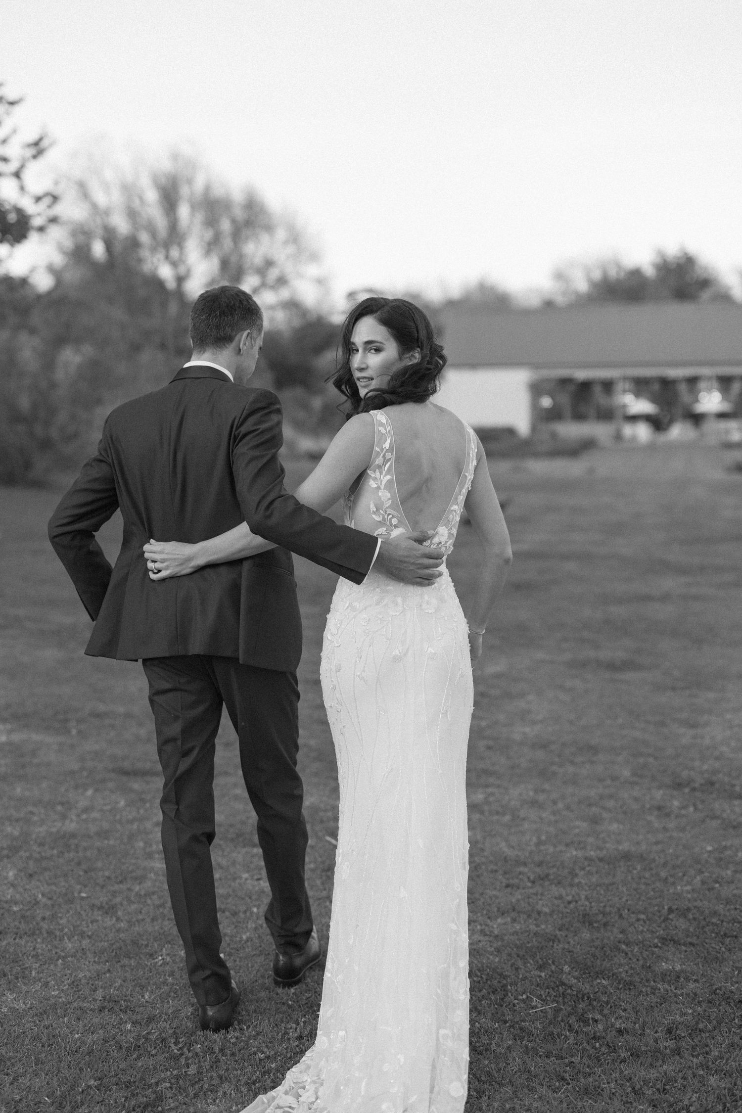 Black and white photo of bride and groom walking outdoors, bride glancing back in elegant lace gown.