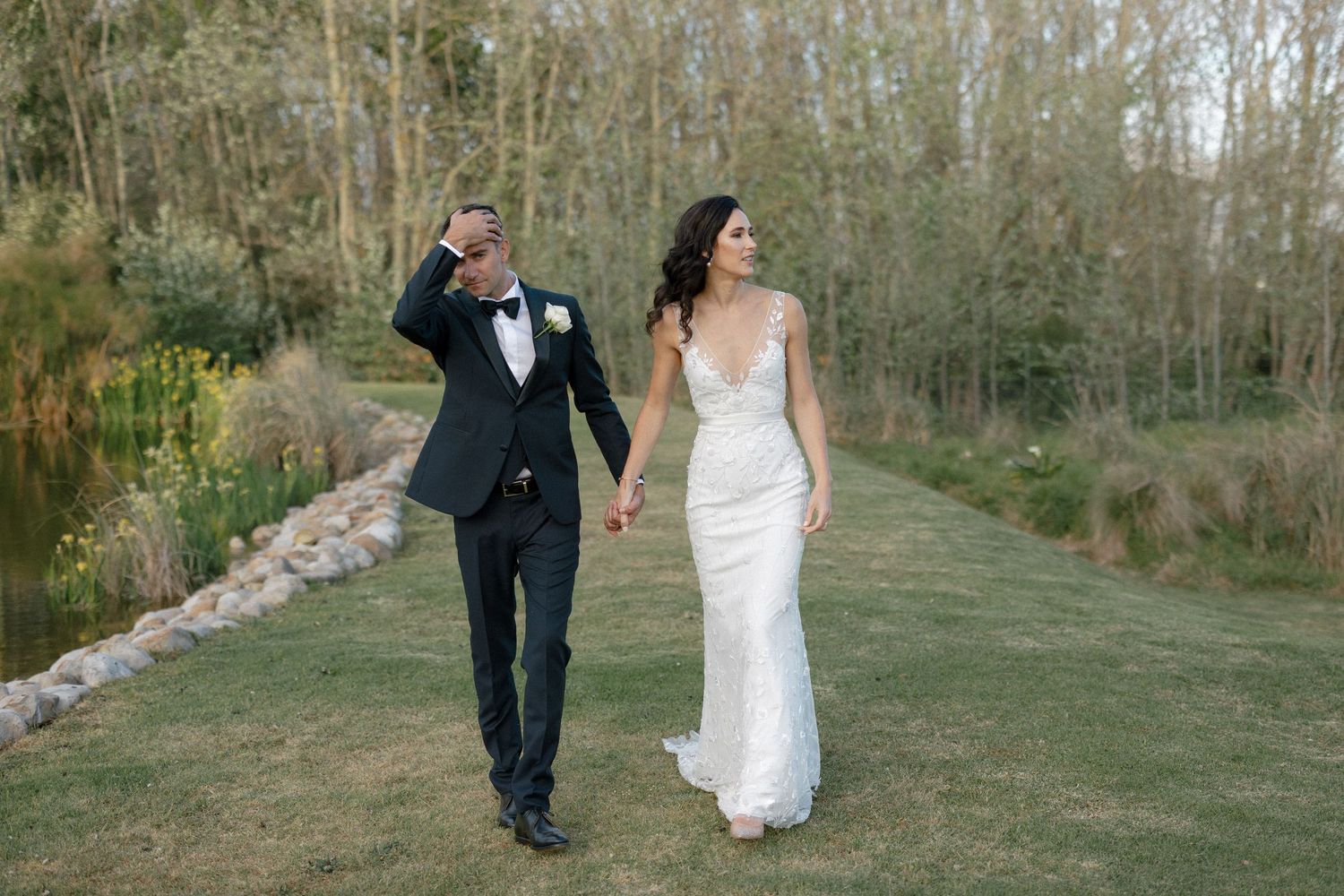 Bride in white lace gown and groom in black tuxedo walking hand in hand through lush garden.