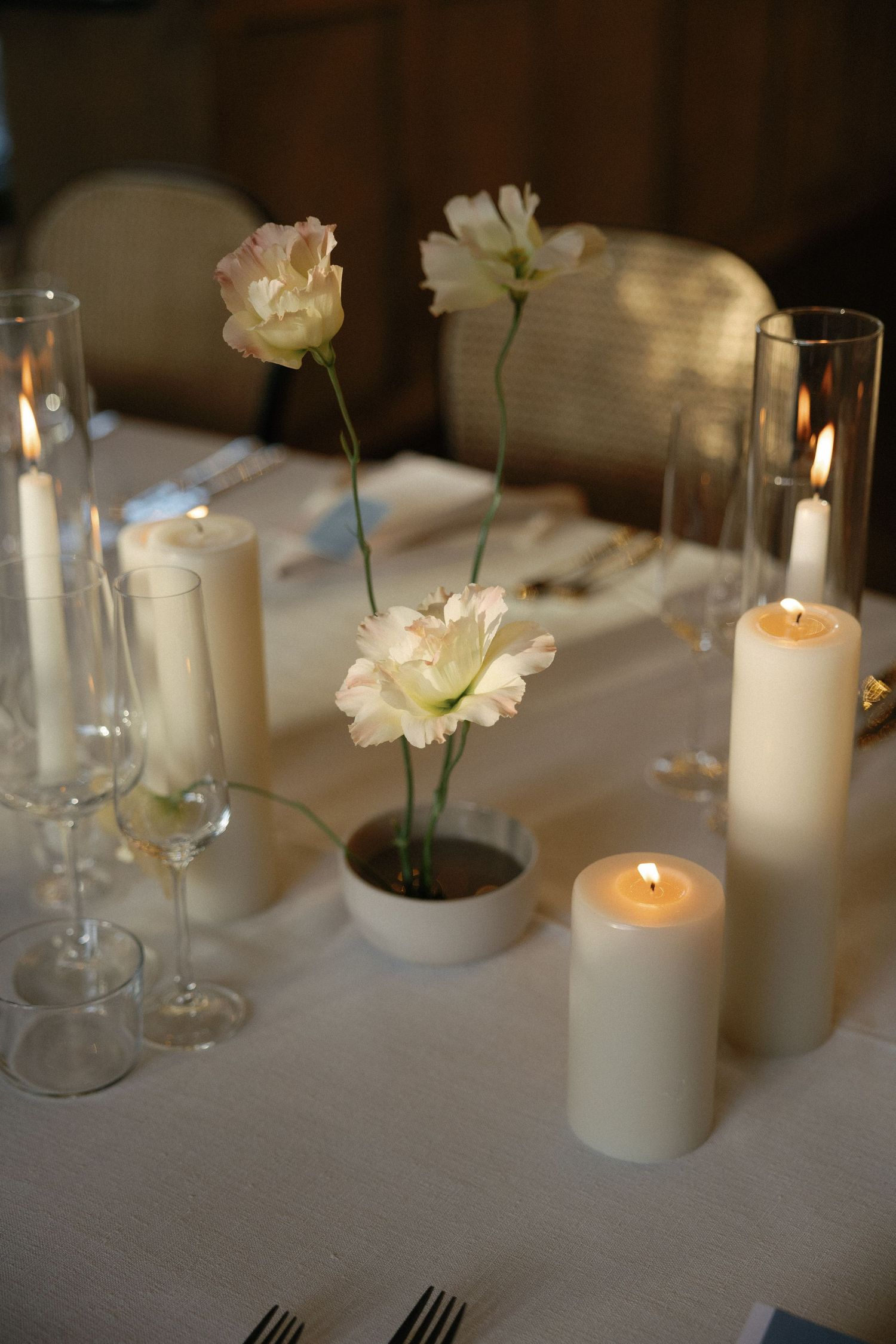 Elegant wedding table setting with lit pillar candles, delicate pink flowers in small vases, and crystal glassware on white linen.
