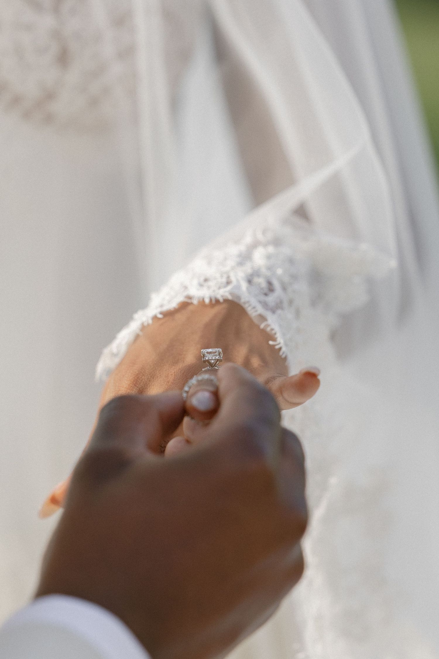 Groom holds brides hand displaying diamond engagement ring against white lace wedding gown.