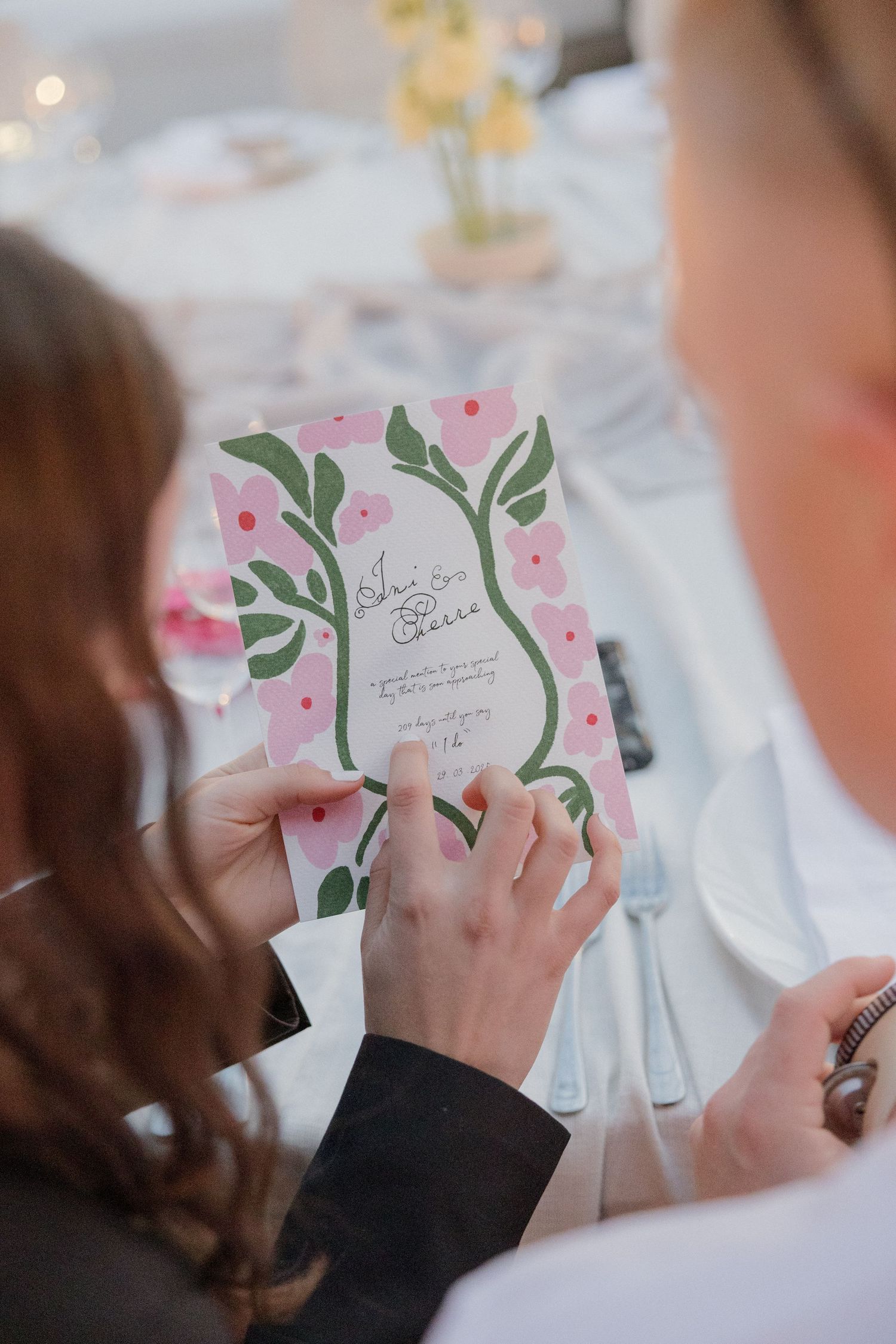 Guests admire a floral pink and green wedding invitation at an elegantly set reception table.