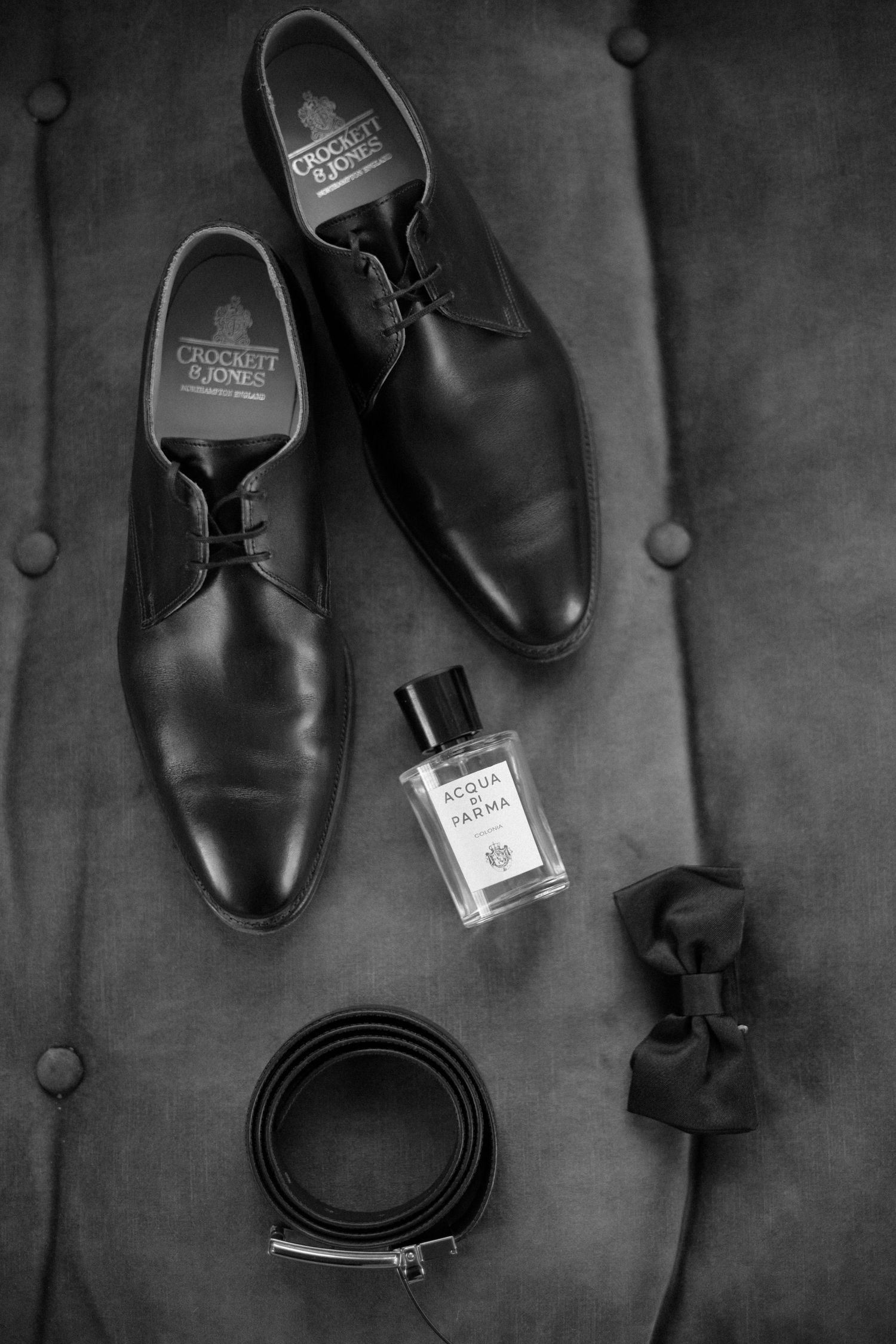 Black and white flat lay of Crockett & Jones dress shoes, Acqua di Parma cologne, belt, and bow tie.