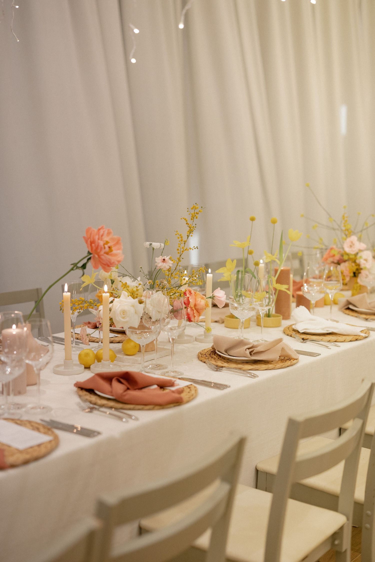 Elegant wedding table setting with pink and yellow floral arrangements, candles, and blush napkins on a white linen tablecloth.