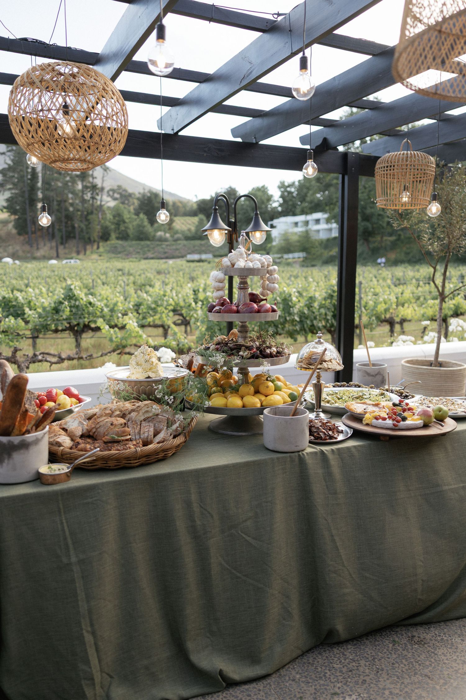 Elegant outdoor buffet table with gourmet food spread, tiered display, and wicker pendant lights overlooking a vineyard.