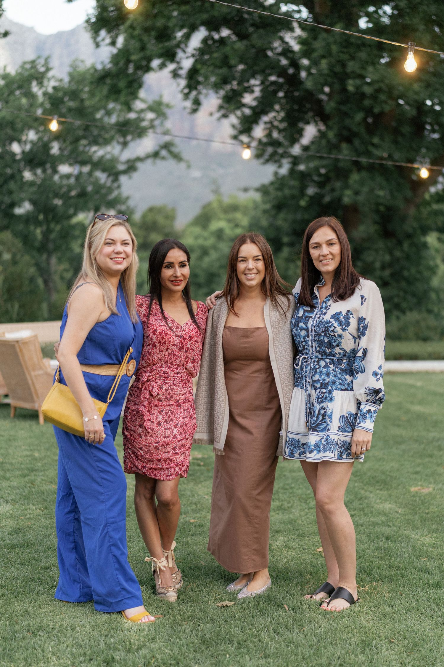 Four women in colorful summer dresses pose together on green lawn with string lights and mountains in background.