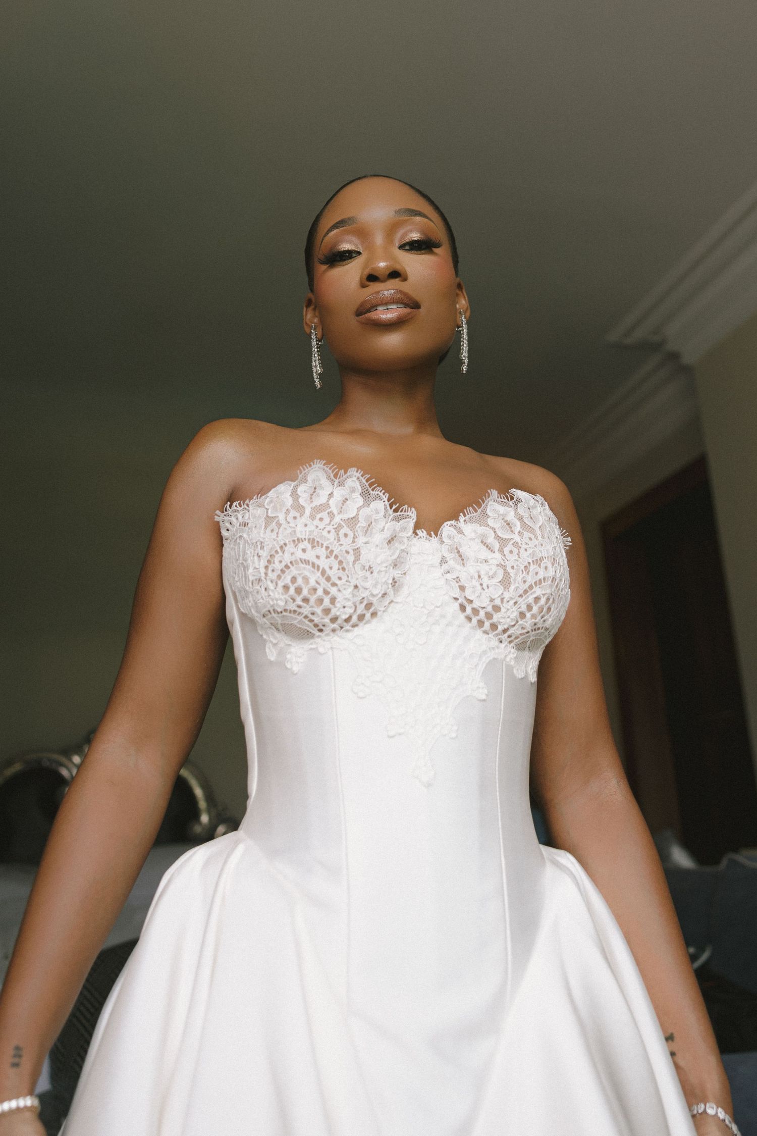 Bride in strapless white lace corset wedding gown with drop earrings, posing elegantly indoors.