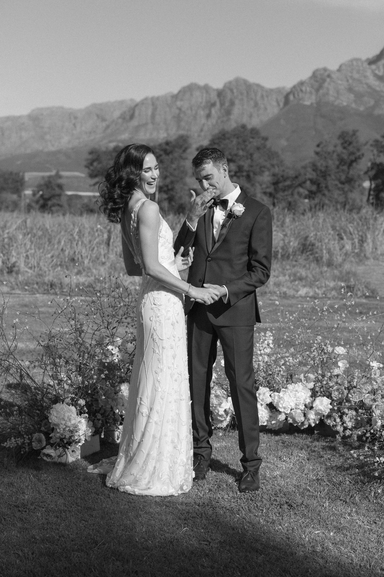 Black and white photo of bride and groom laughing during outdoor wedding ceremony with mountain backdrop.