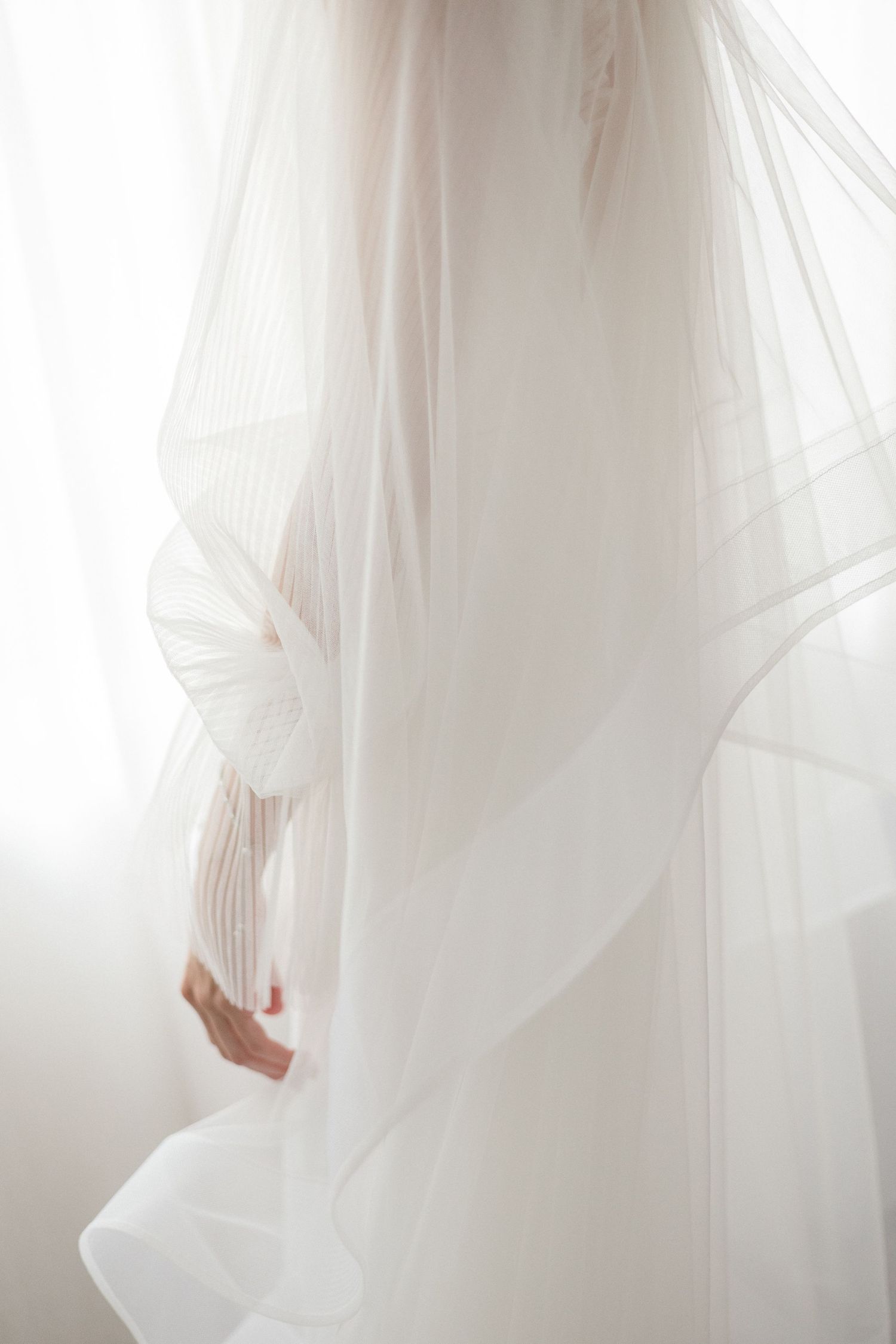A bride draped in a flowing white tulle wedding veil, with soft light filtering through sheer curtains.