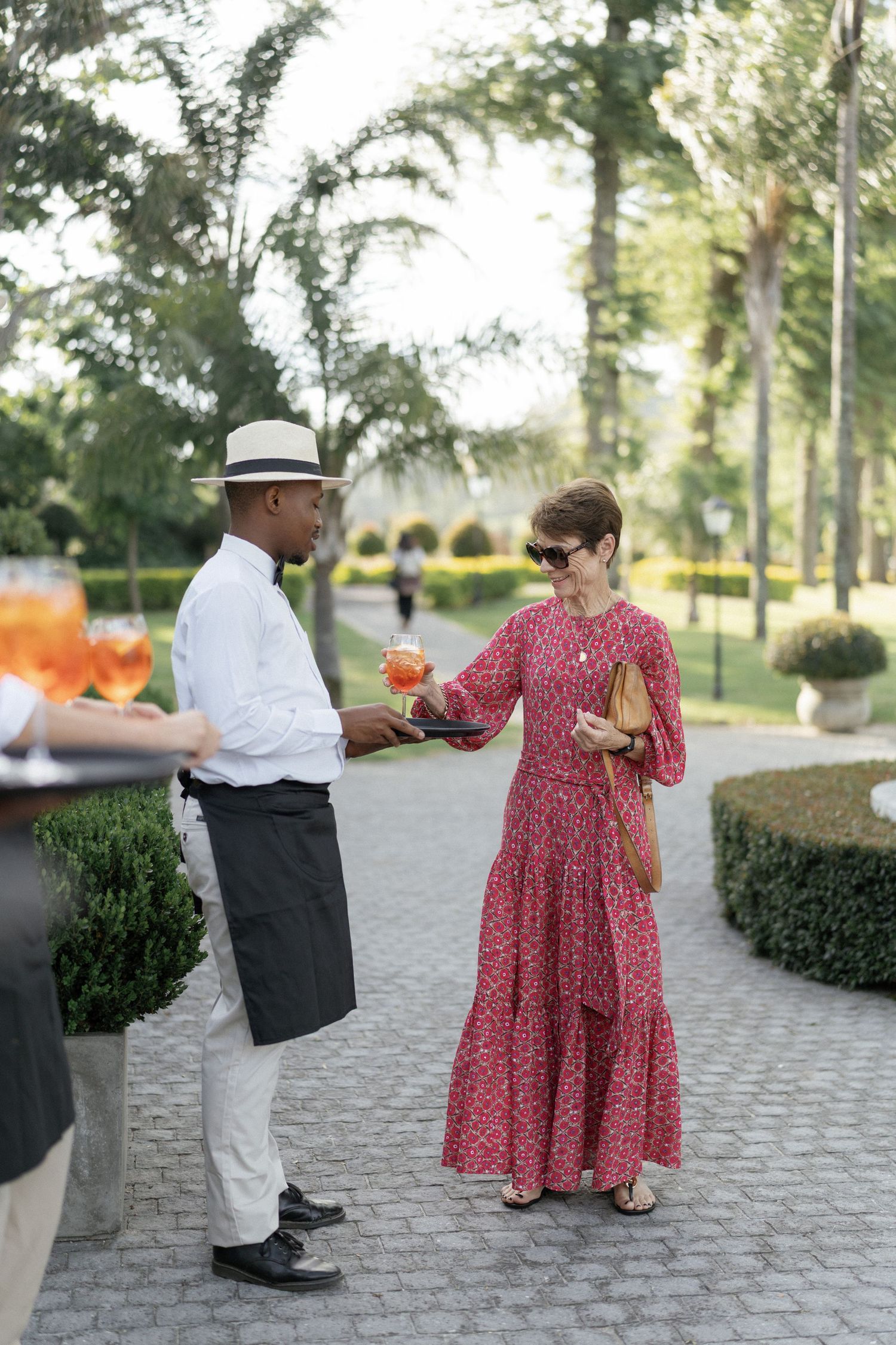 A waiter in a white shirt and hat serves a cocktail to a woman in a red floral dress at an outdoor garden event.