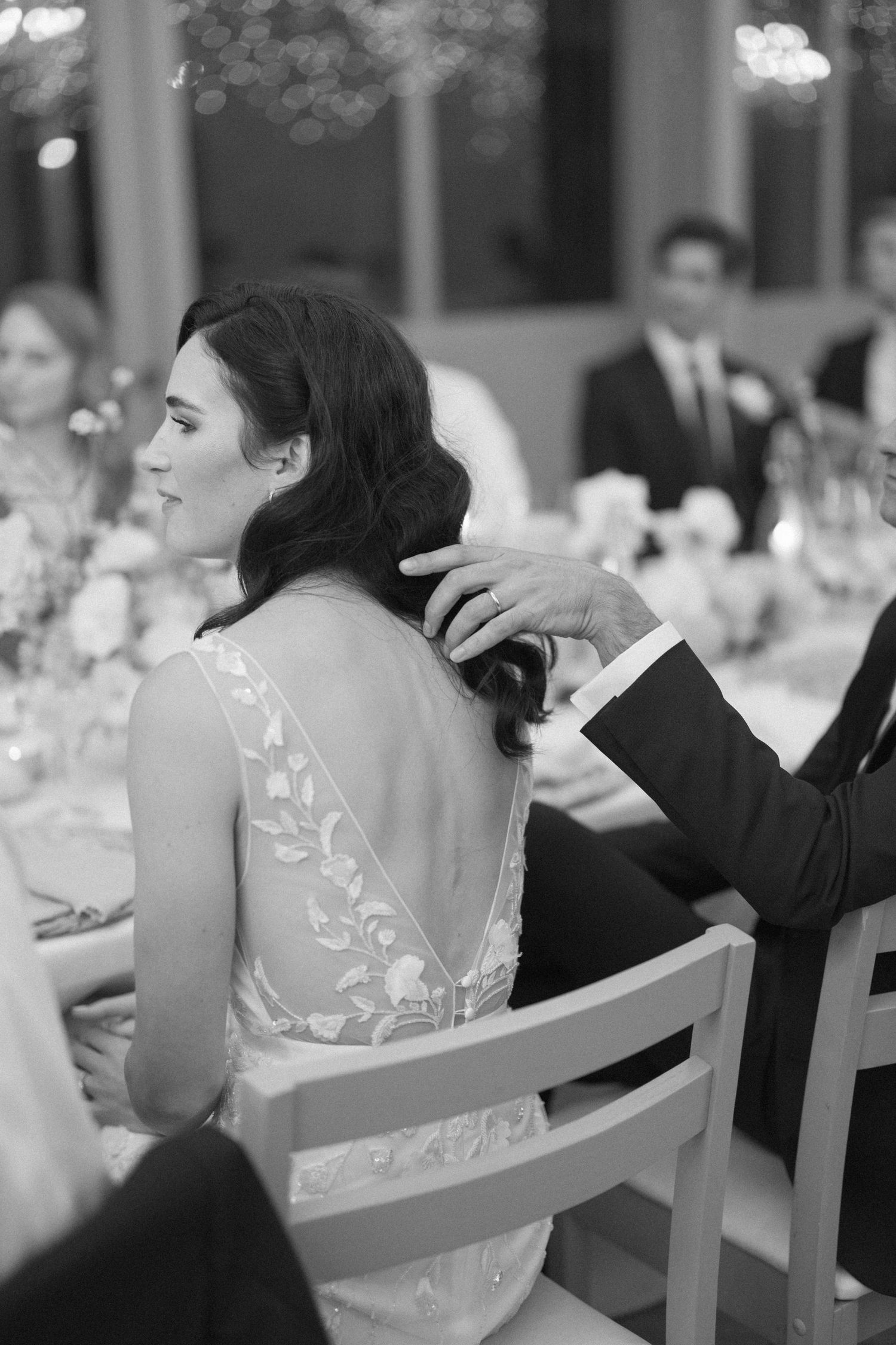 Bride in backless lace wedding gown receives tender touch on back during wedding reception.