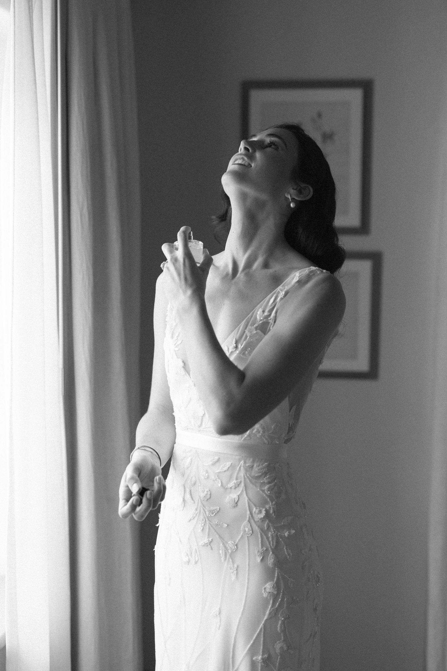 Black and white photo of a radiant bride in a beaded gown laughing joyfully while applying perfume near a window.