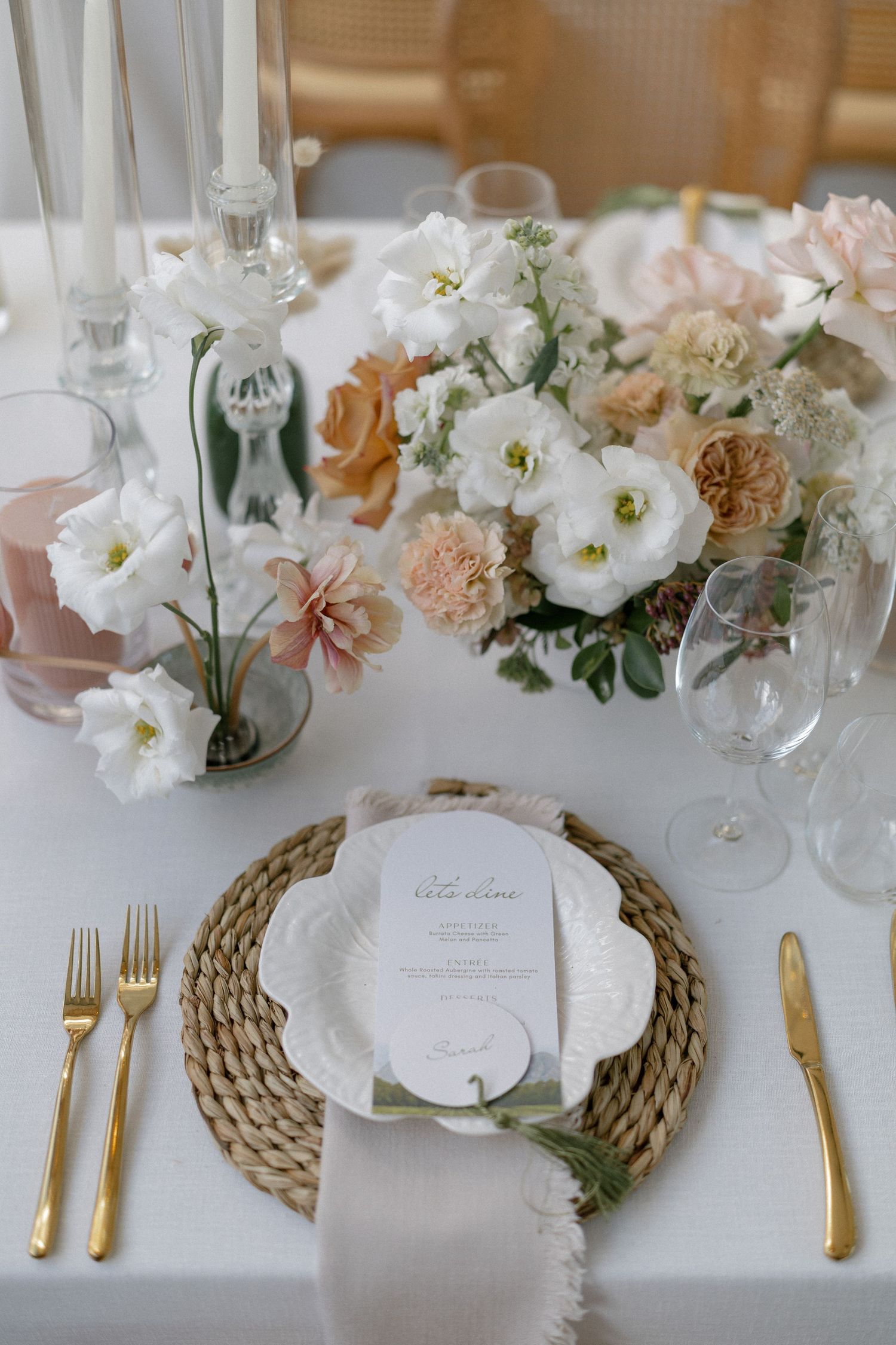 Elegant wedding table setting with gold cutlery, woven charger, white floral centerpiece, and soft peach blooms.