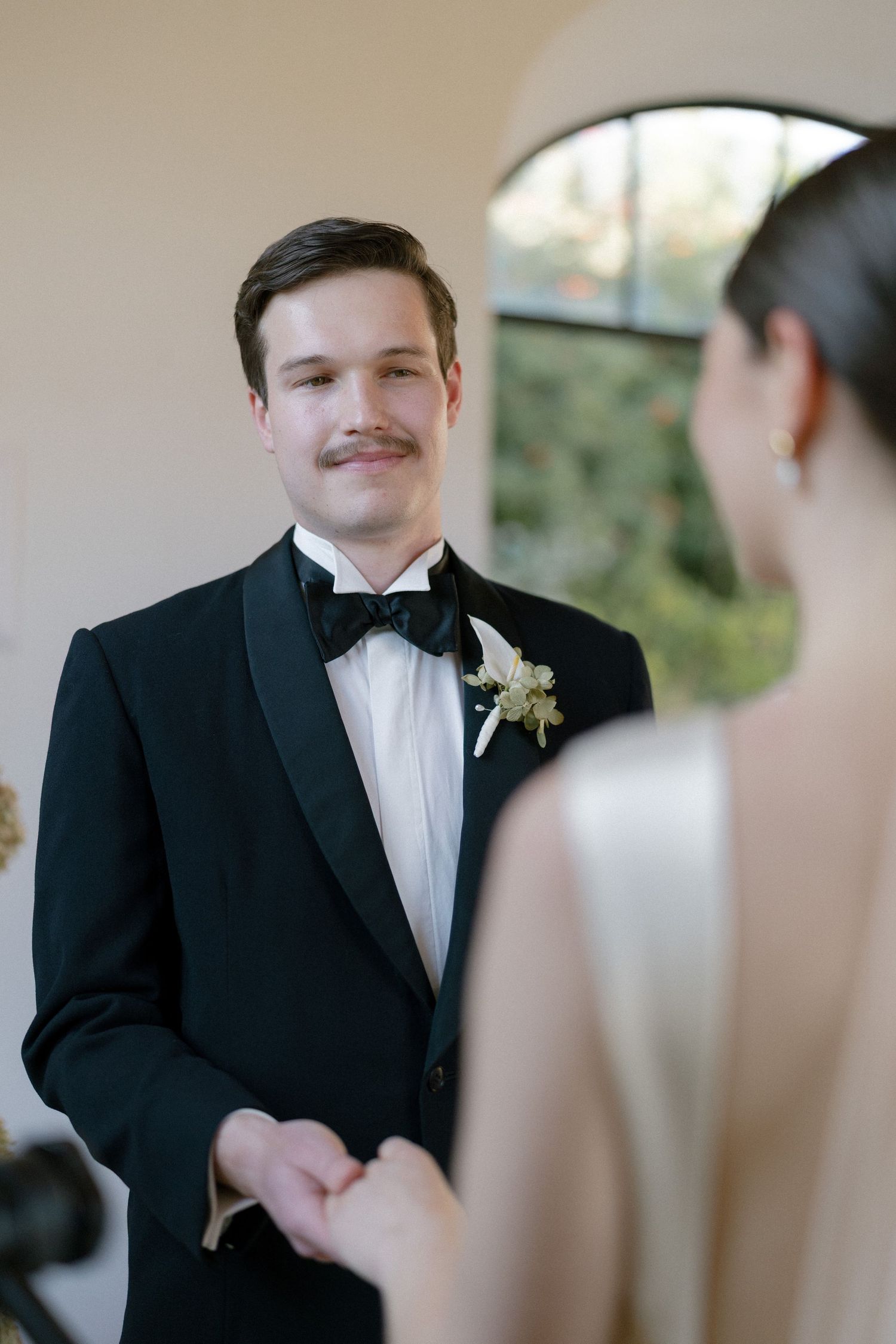 Groom in black tuxedo with boutonniere smiles at bride during wedding ceremony.