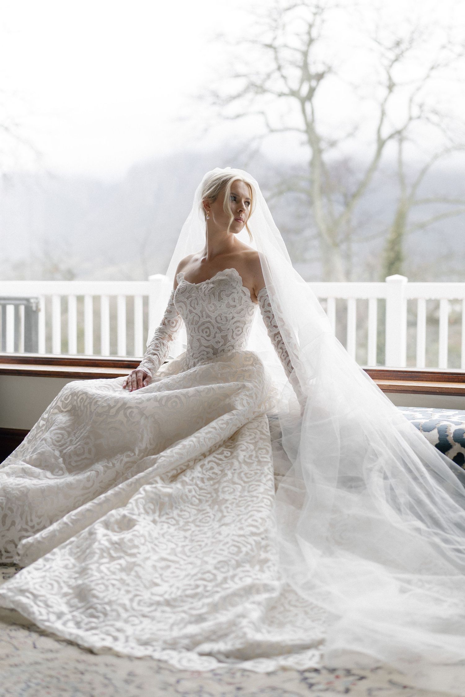 Bride in elegant lace wedding gown with long veil seated by window with misty mountain backdrop.