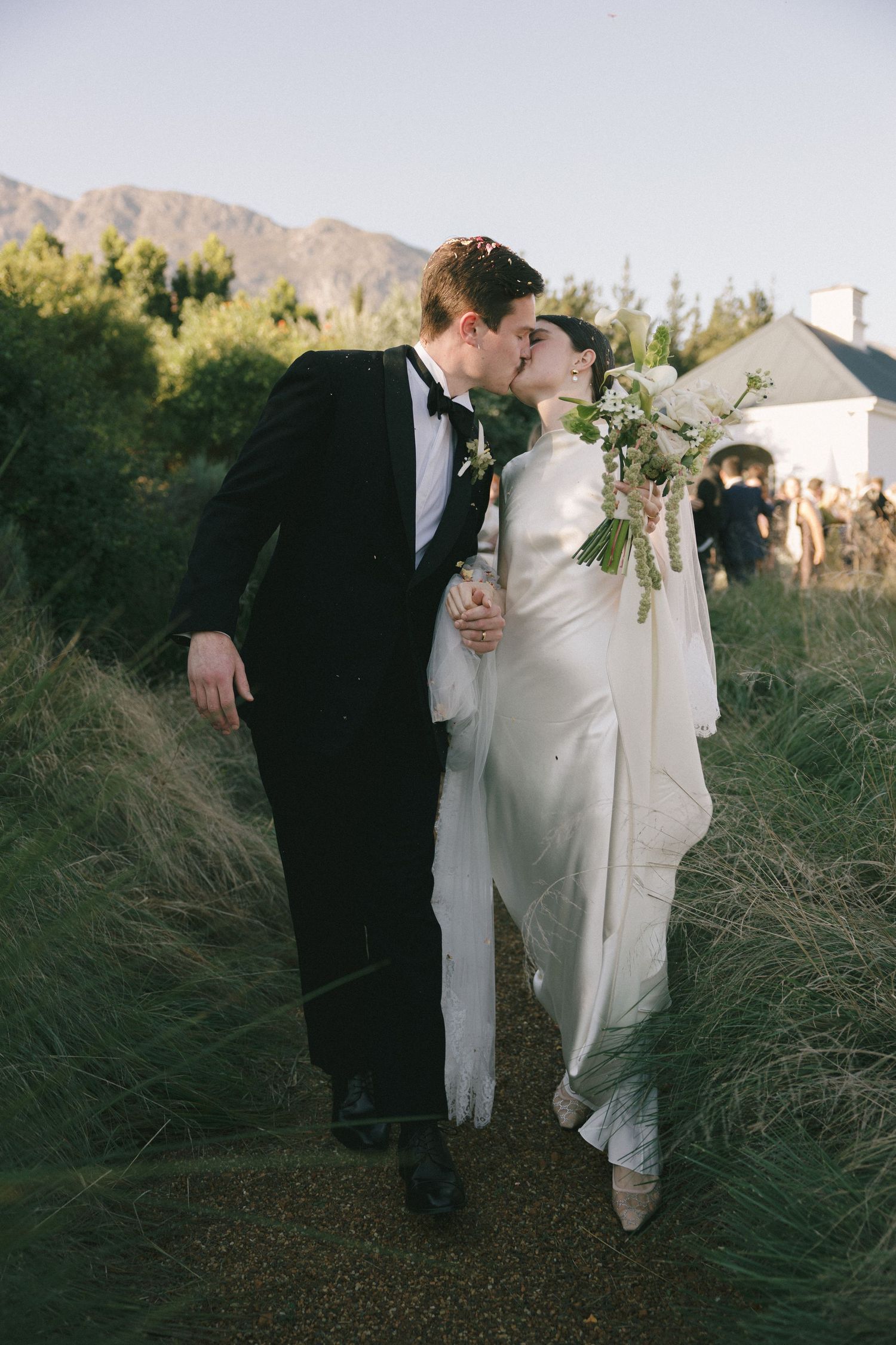Bride and groom share a kiss while walking through tall grass, she holds a white bouquet, mountains visible in background.