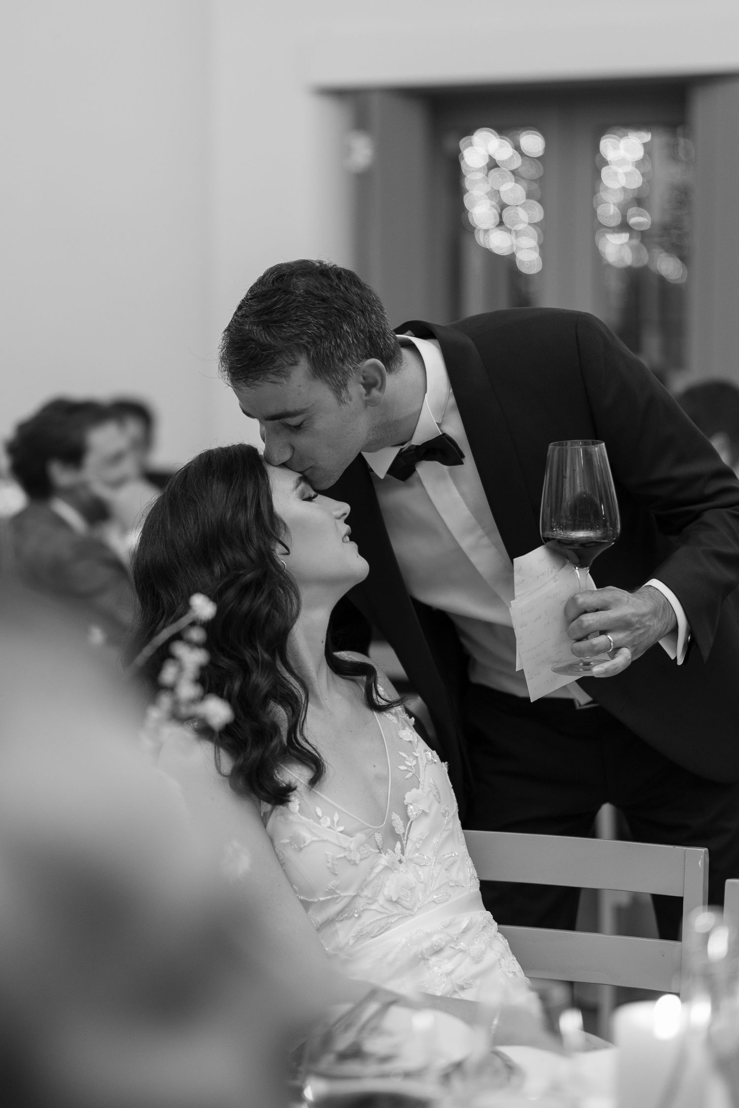 Groom kisses seated bride while holding wine glass at wedding reception in black and white.