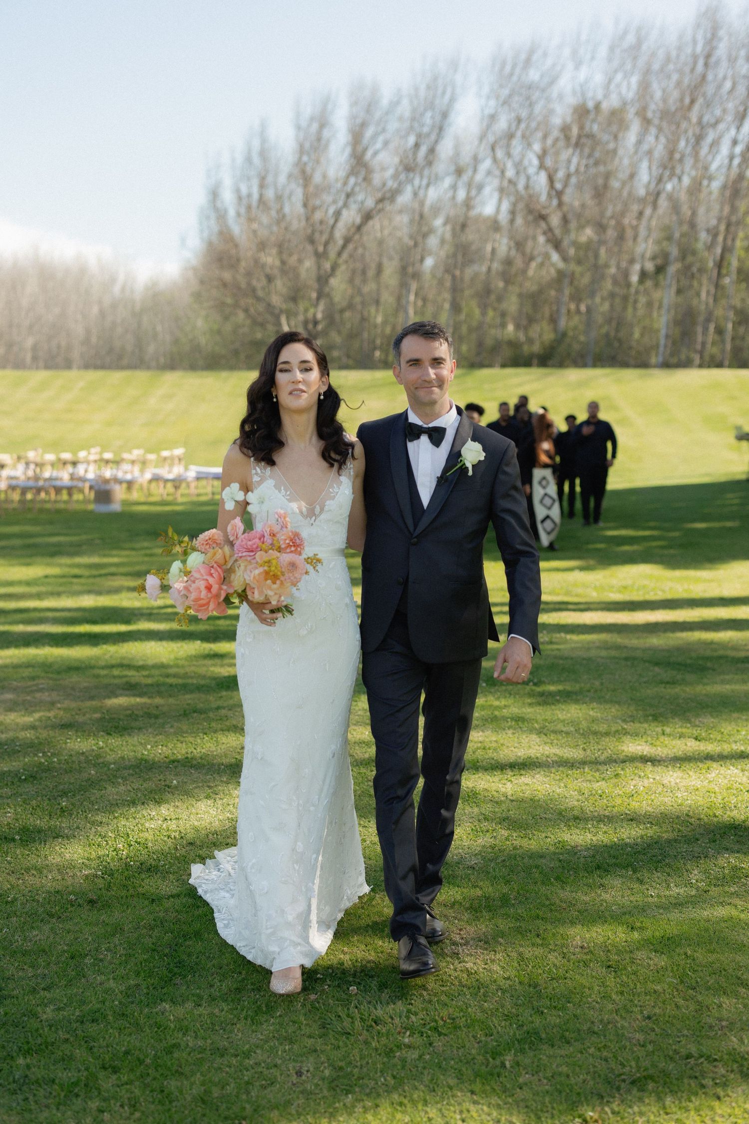 Bride and groom walking down grassy aisle after outdoor wedding ceremony, bride holding pink bouquet.