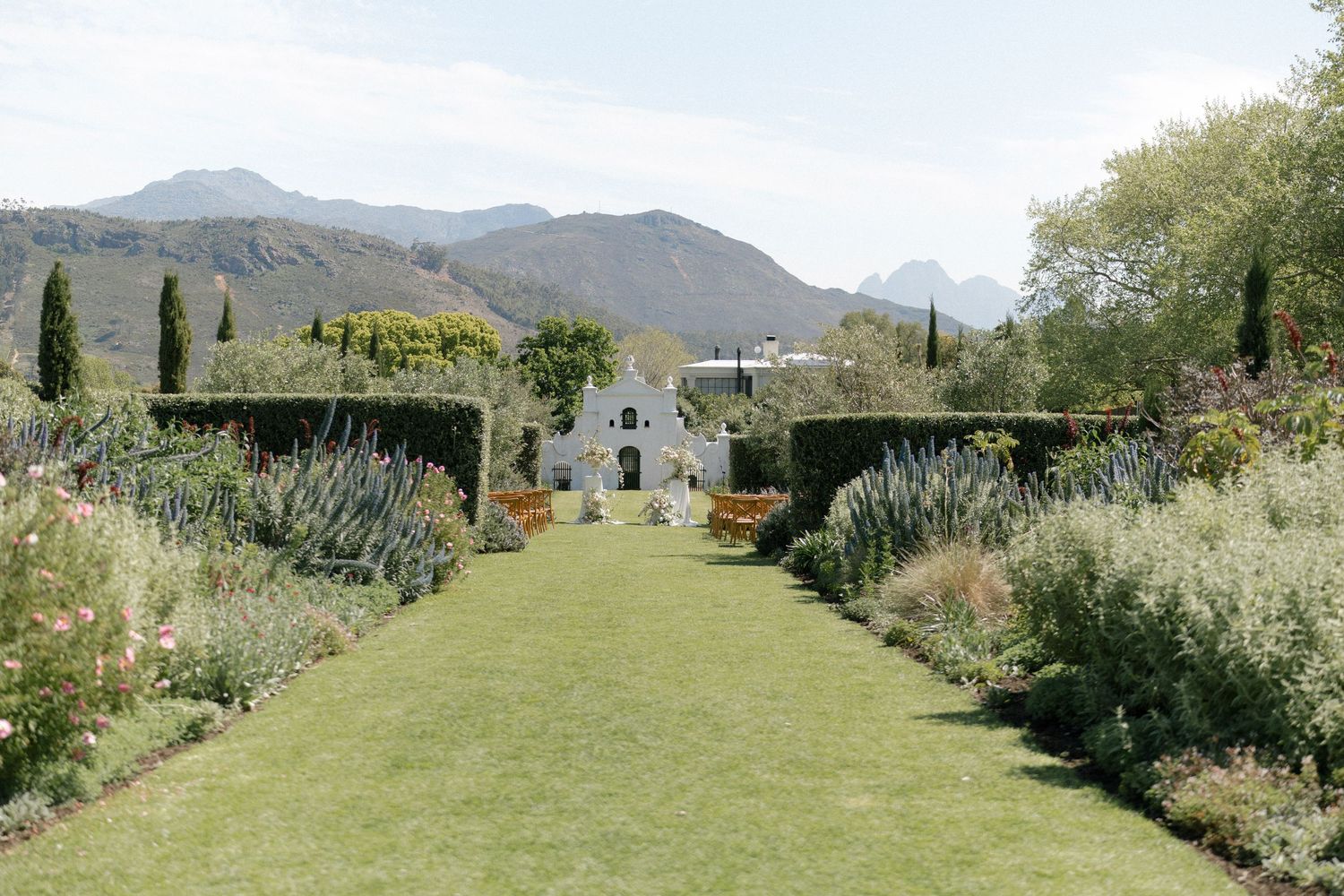 Lush formal garden with green lawn, colorful borders, white Cape Dutch building, and mountain backdrop under clear skies.