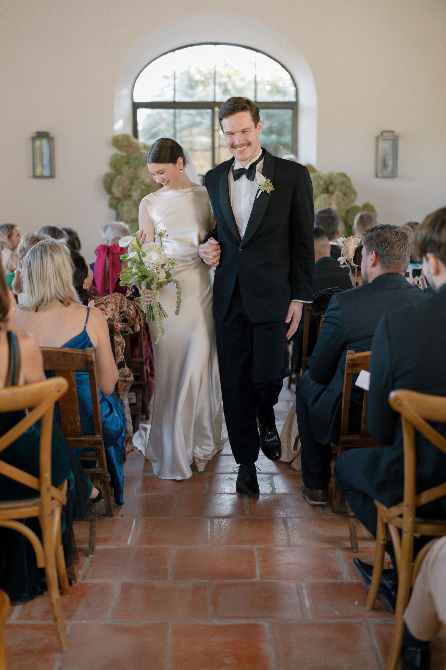 Newlyweds walking down the aisle together smiling after wedding ceremony in elegant chapel with terracotta floors.