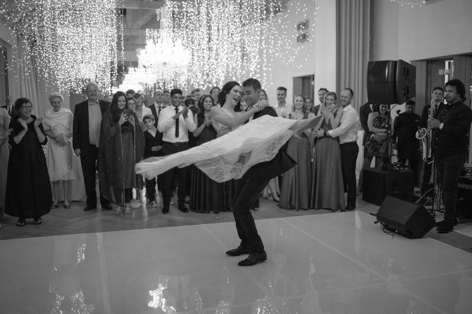 Groom lifts bride in flowing white gown during first dance surrounded by applauding guests.