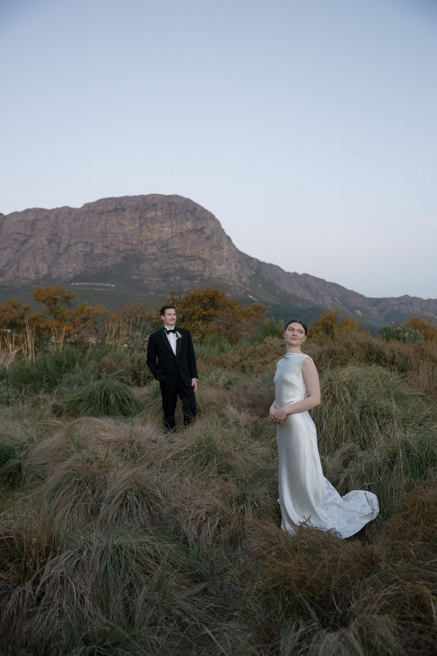 Bride and groom stand apart in tall grass with dramatic mountain backdrop at dusk in South Africa.